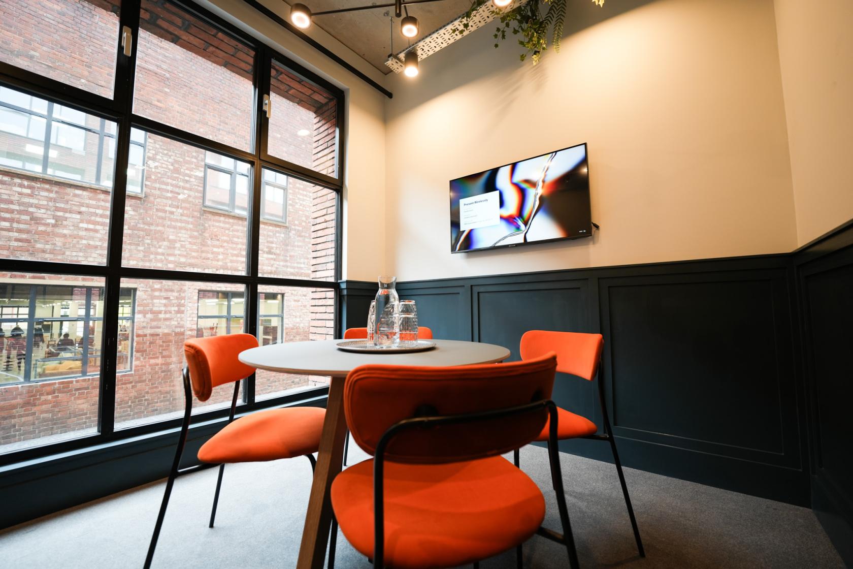 A meeting room with large windows, orange chairs, a round table, and a wall-mounted screen at Colony One Silk Street.