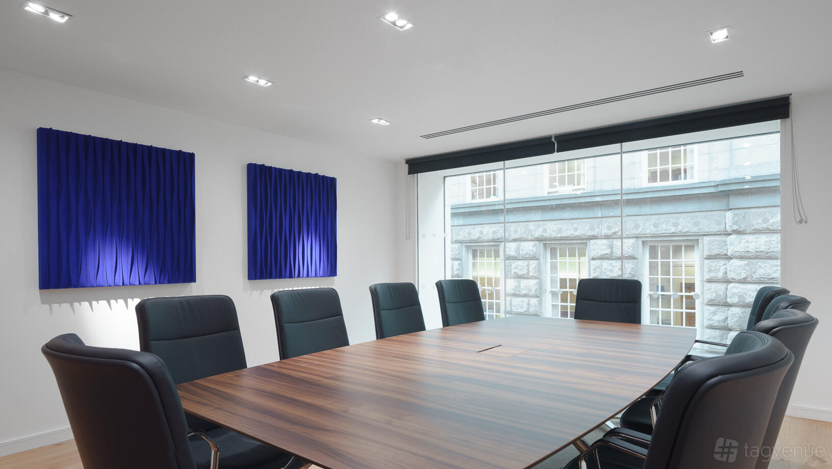 A boardroom with a large wooden table, black leather chairs, floor-to-ceiling windows, and blue acoustic panels at Viridis Offices.