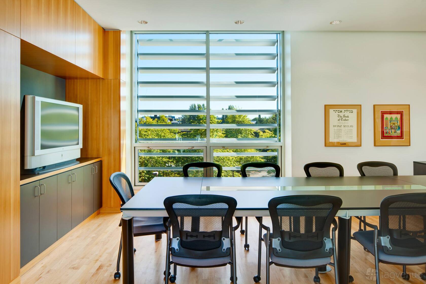 A boardroom with a long glass-top table, black mesh chairs, and large windows with tree views at Hillel BC.