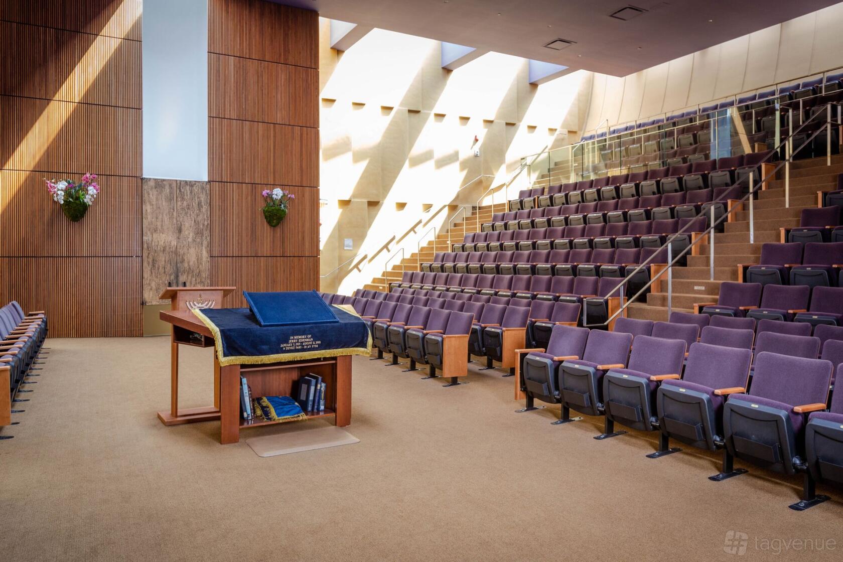 An auditorium with tiered seating, wood panel walls, and a lectern under skylights at Congregation Beth Sholom.