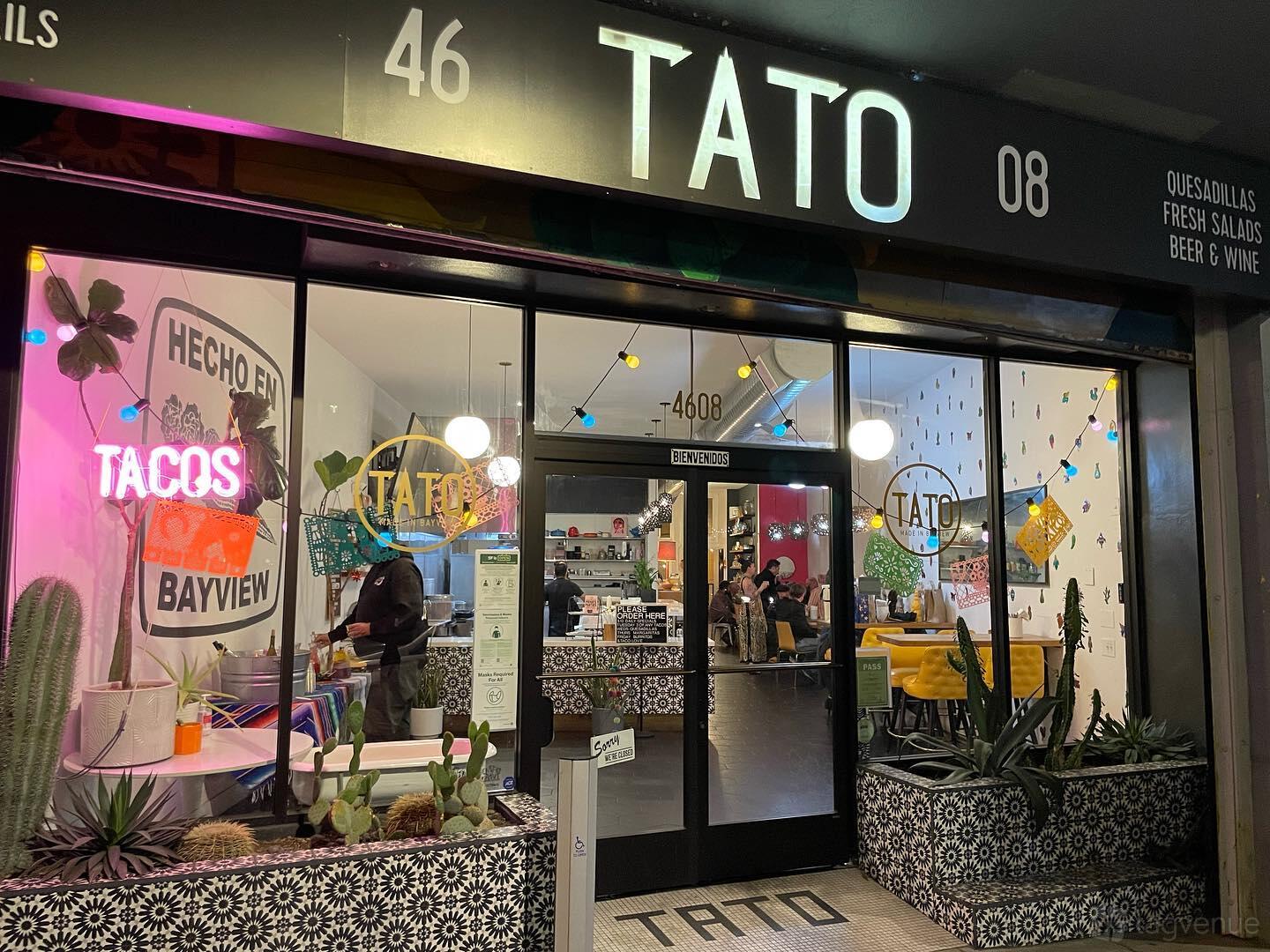 A restaurant with large glass windows, patterned tile entrance, and neon taco sign at Tato.