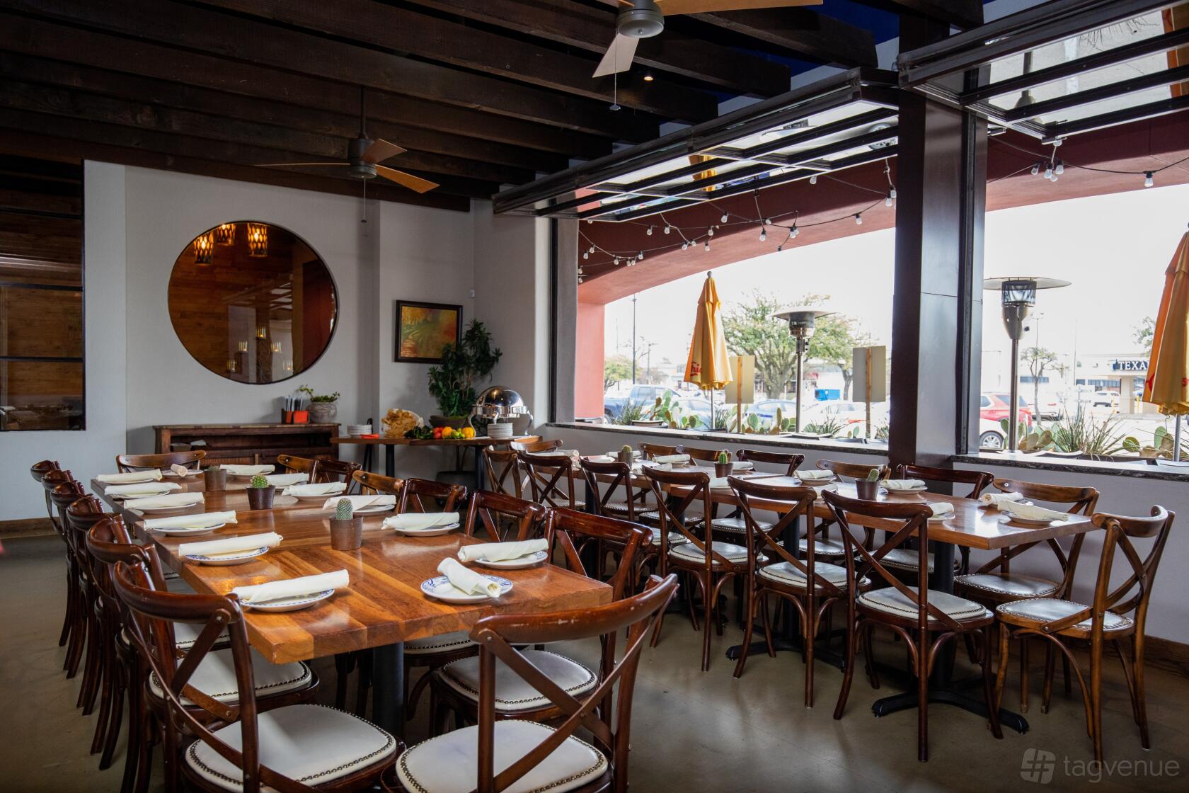 An event space in a restaurant with long wooden tables, cross-back chairs, large windows, and ceiling fans at Meso Maya - Preston Forest.