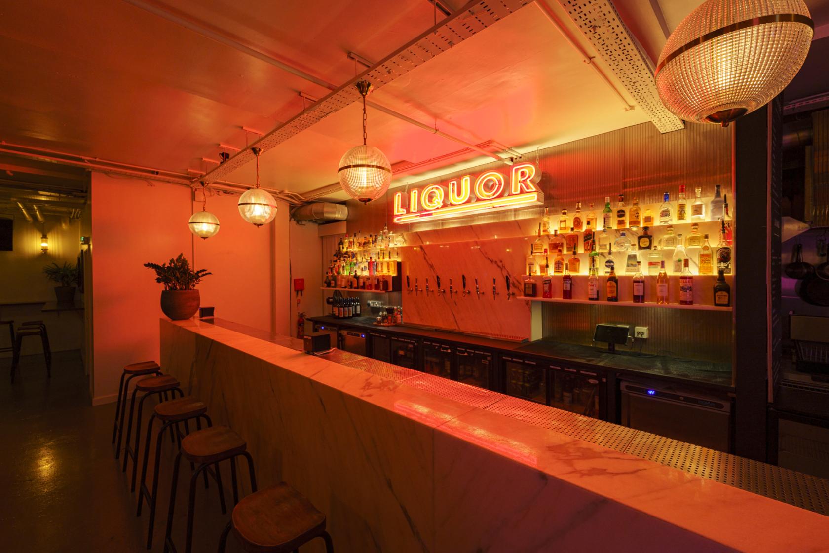 A cocktail bar with a marble counter, backlit shelves of spirits, and a neon ‘Liquor’ sign at NT's Loft.