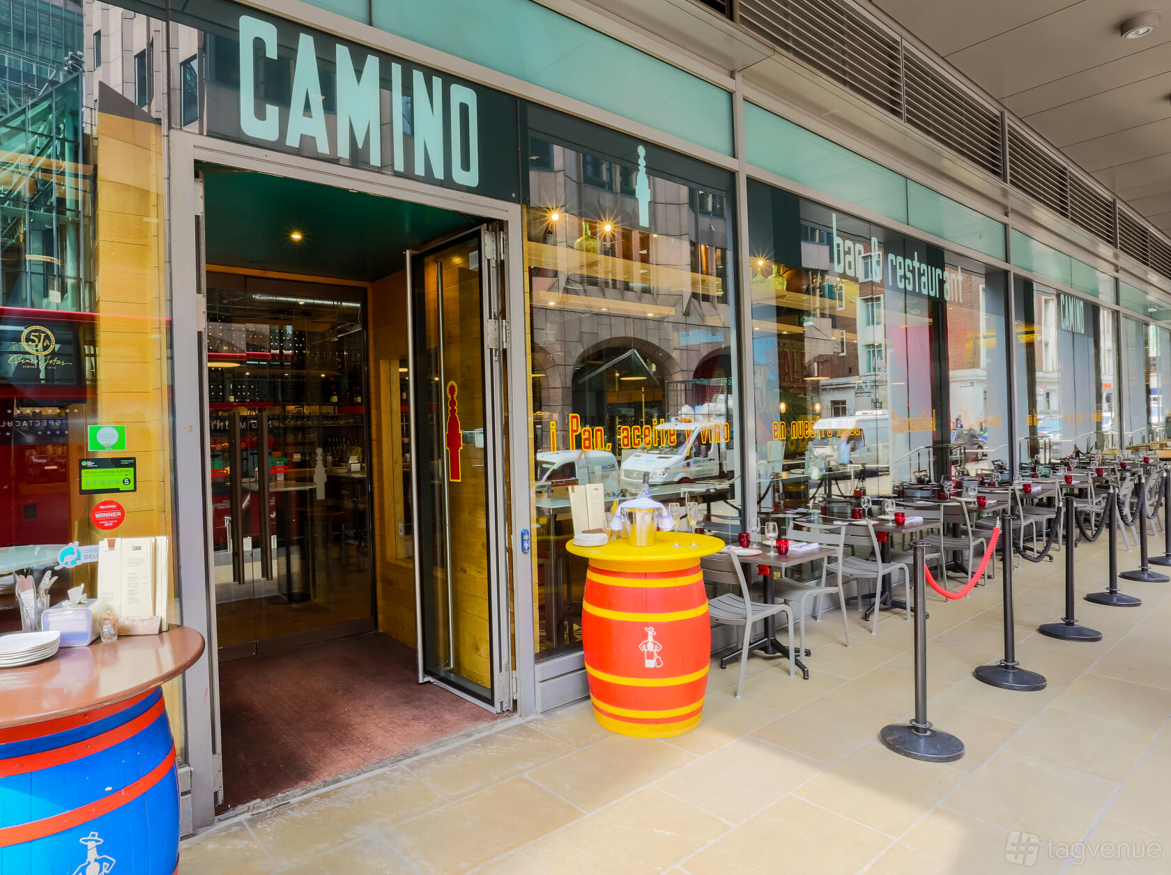 A restaurant with outdoor seating, glass facade, and bright barrel tables at Camino Monument.