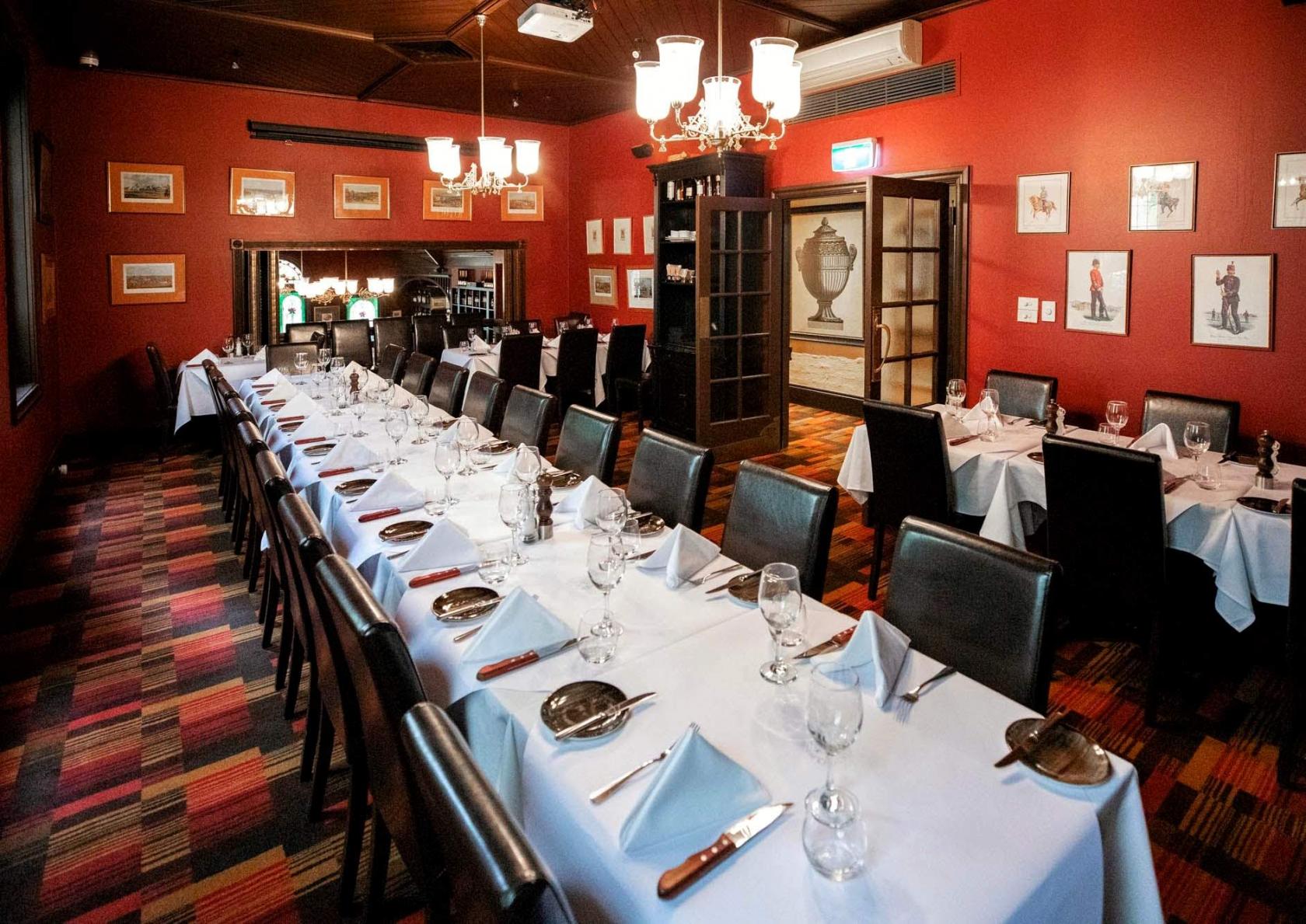 A dining room with two long tables set with white linens, glassware, and chandeliers at The Mitre Steakhouse.