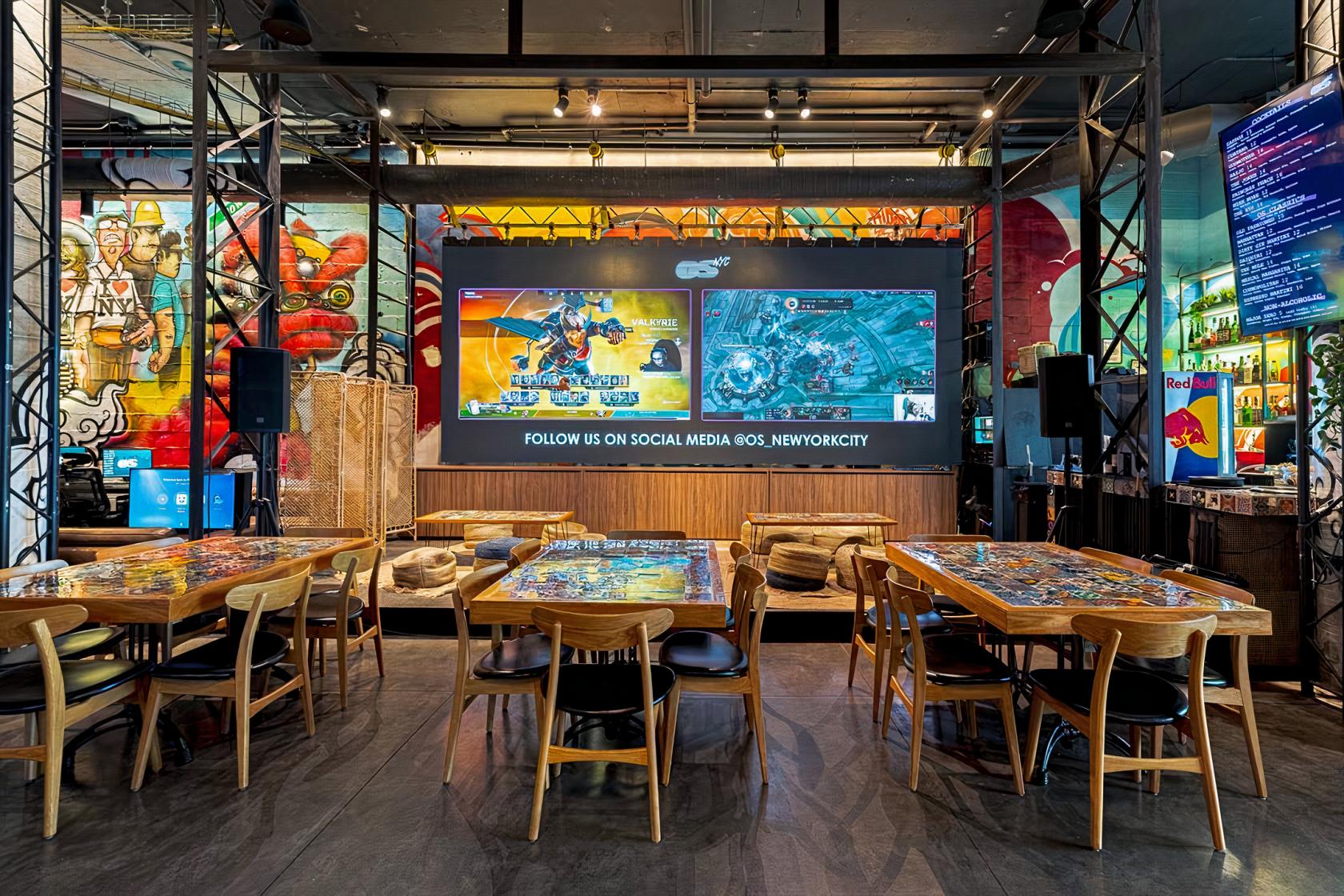 A bar event area with arcade-inspired wall art, large gaming screens, and wooden tables with gaming graphics at OS NYC.