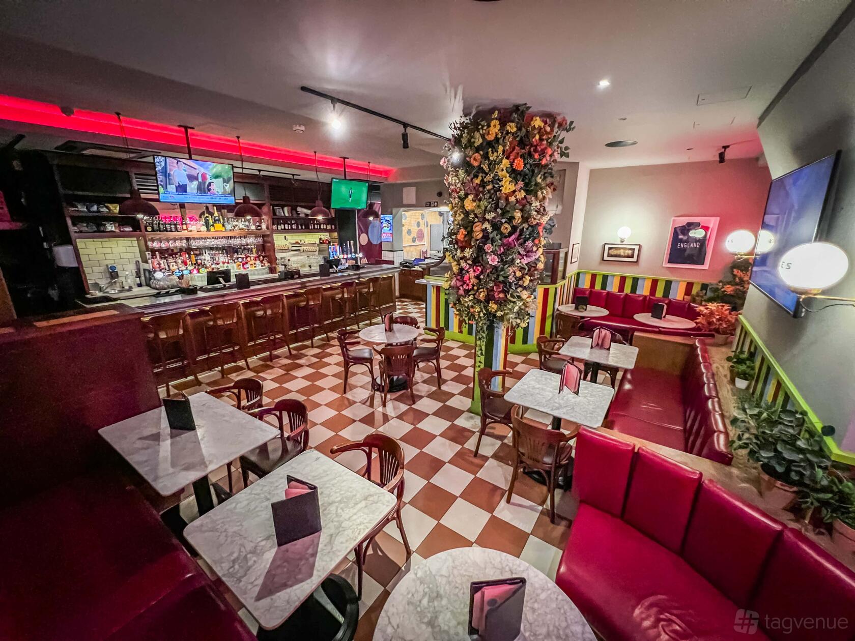 A bar with a checkered floor, marble tables, red booth seating, and a flower-covered pillar at Sixes Social Cricket - Fitzrovia.