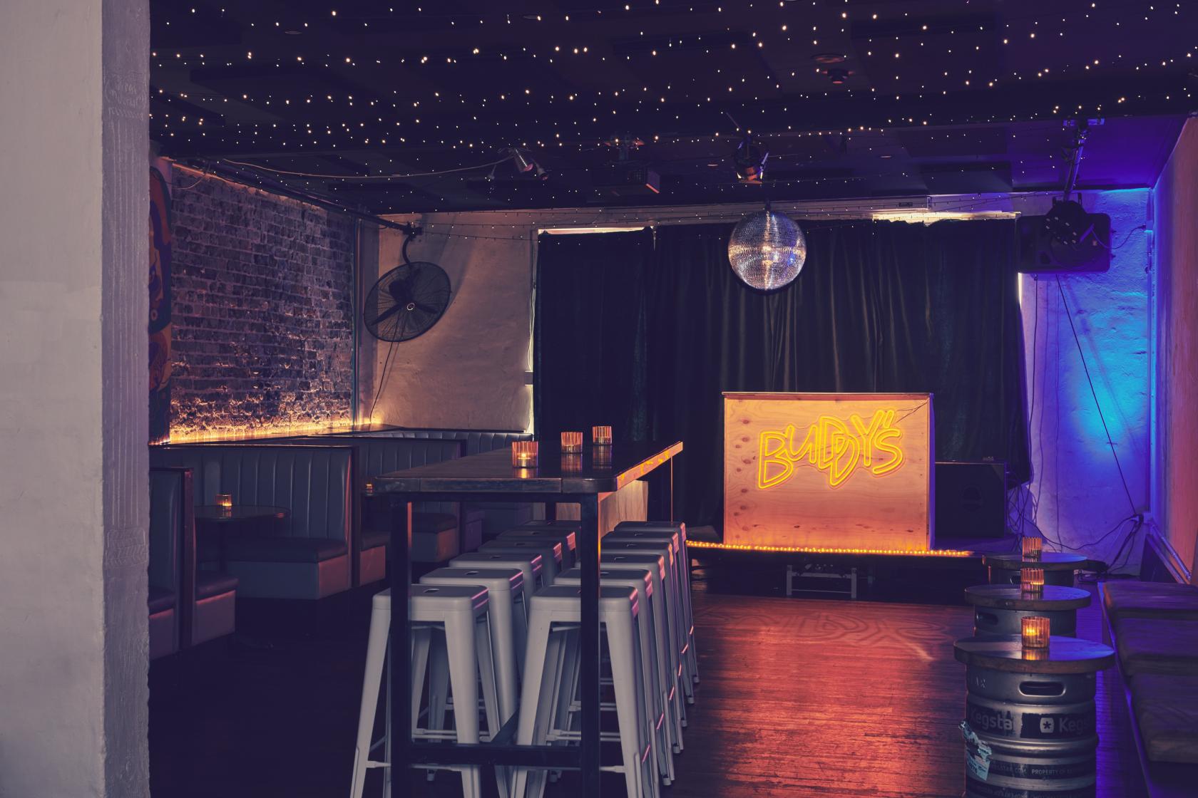 An upstairs event space with exposed brick walls, string lights, and a neon Buddy's sign at Buddy's Bar Newtown.