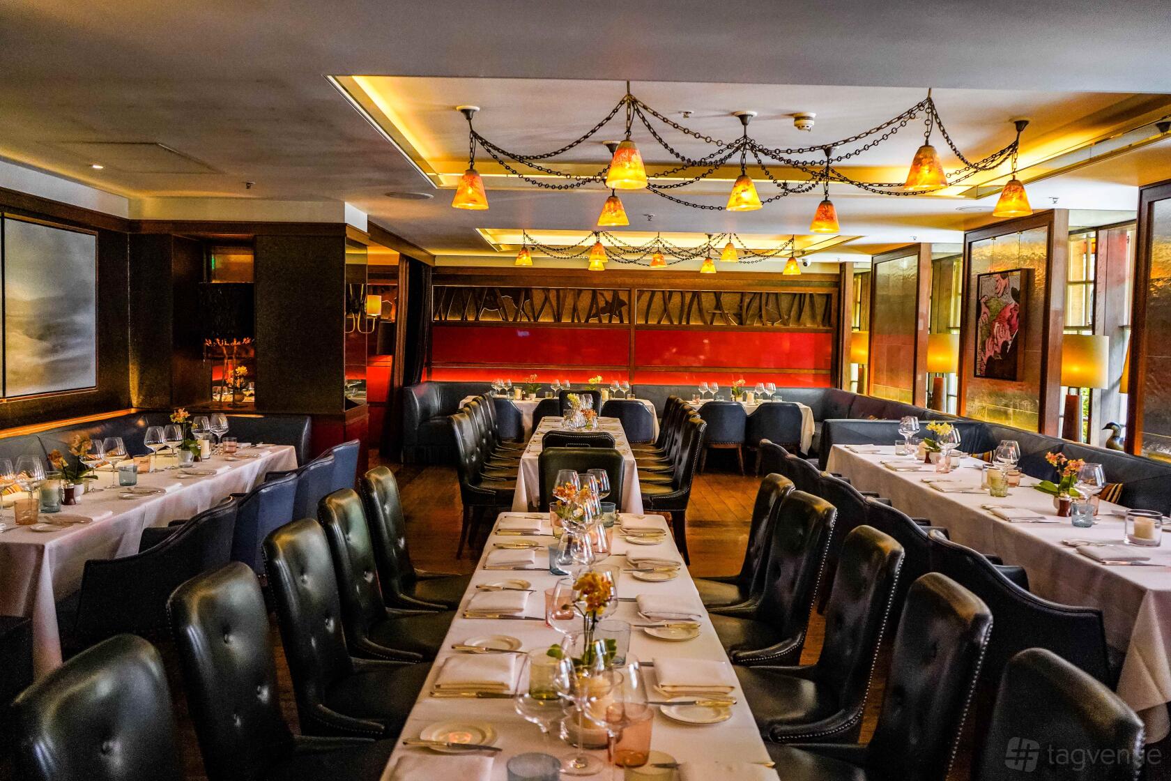 A restaurant dining room with long tables set with white linens, dark leather chairs, and warm pendant lighting at Corrigan's Mayfair.