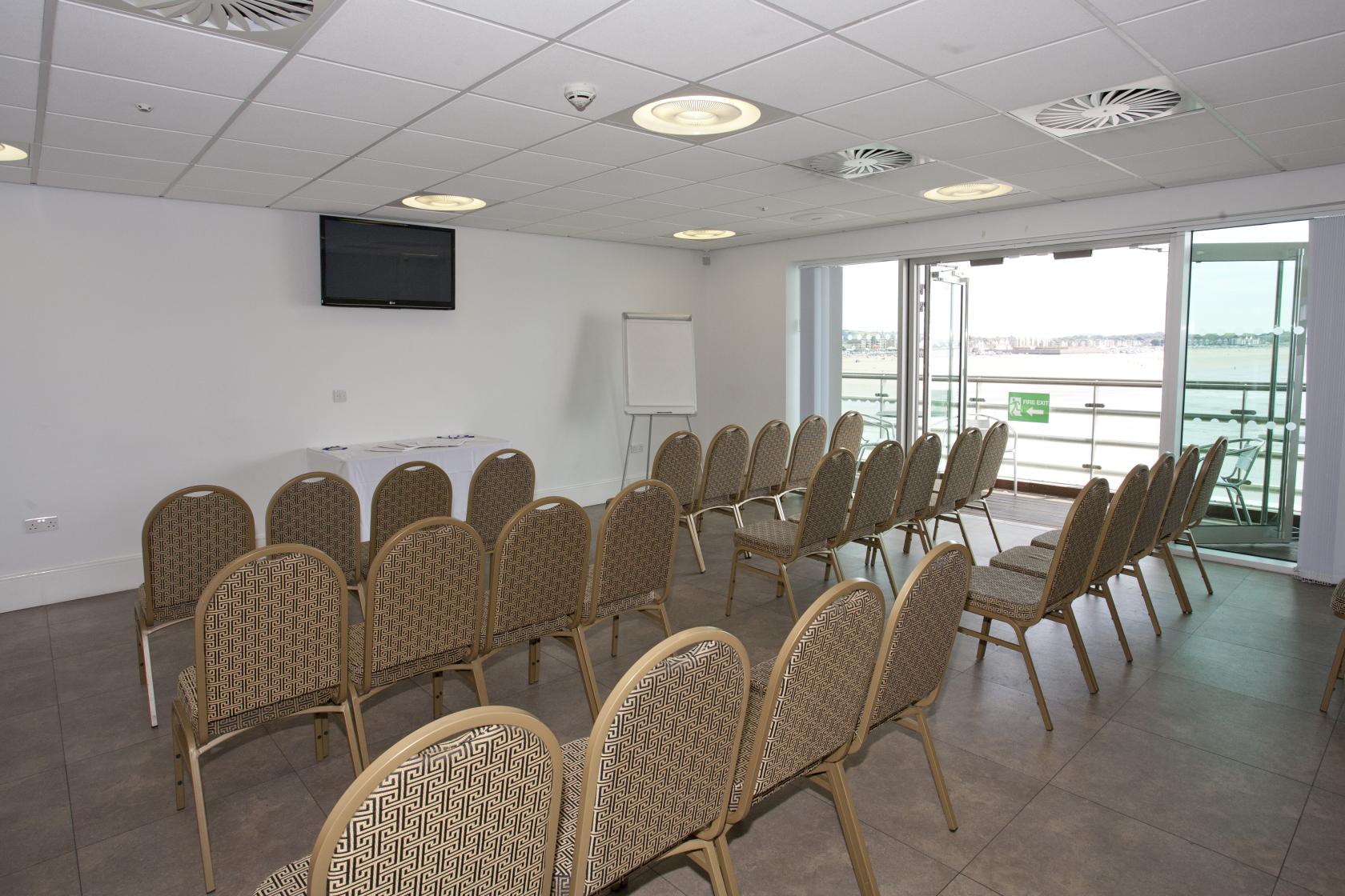 A boardroom with rows of patterned chairs, a wall-mounted TV, and balcony access overlooking the water at The Grand Pier.