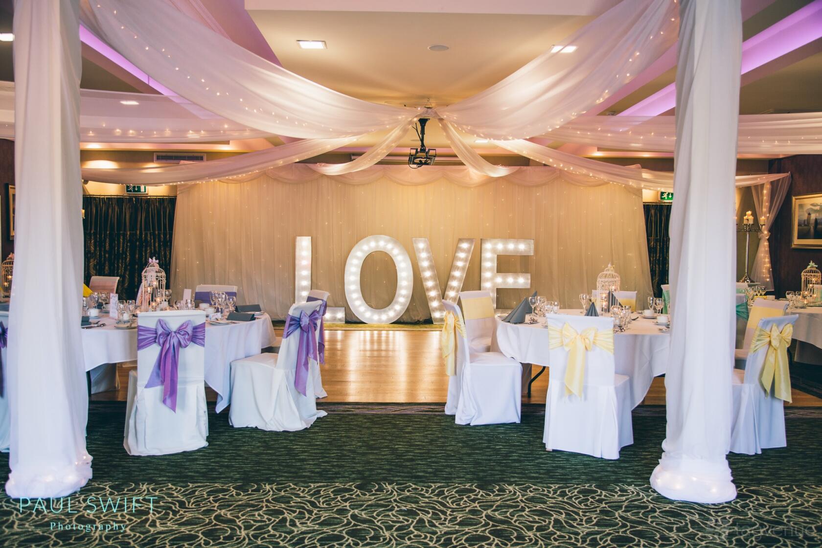 An event venue with draped ceilings, large illuminated 'LOVE' letters, and round tables with pastel chair sashes at Wyrebank.