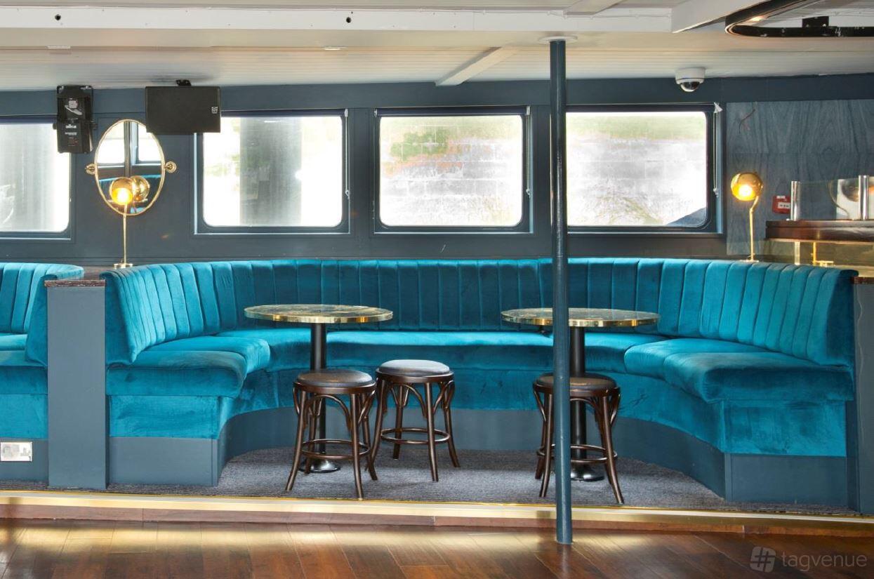 An event space on a boat with blue velvet banquette seating, round tables, and brass floor lamps at The Tattershall Castle.
