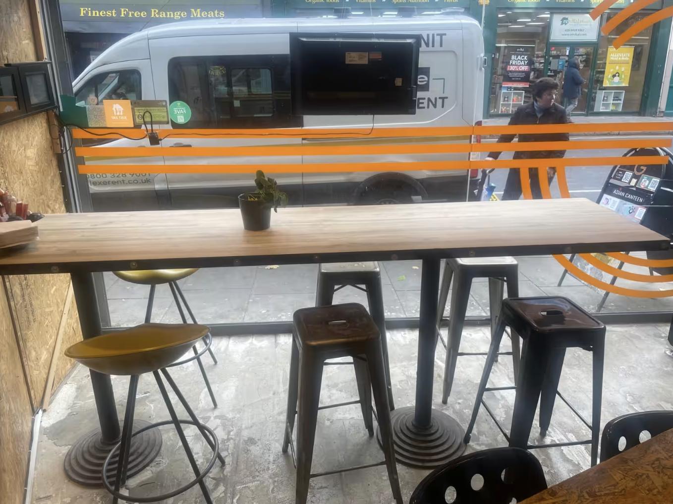 A restaurant dining area with a long wooden high-top table, metal stools, and a small potted plant at G Asian Canteen.