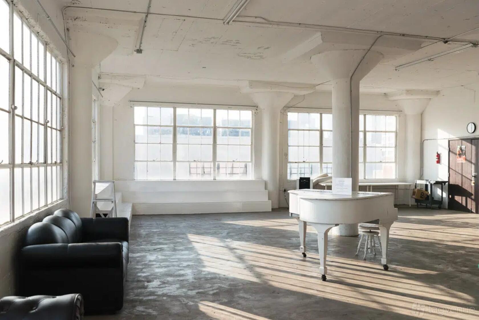 A photo studio with large industrial windows, white steps, a white grand piano, and exposed concrete floors at Art Studios.