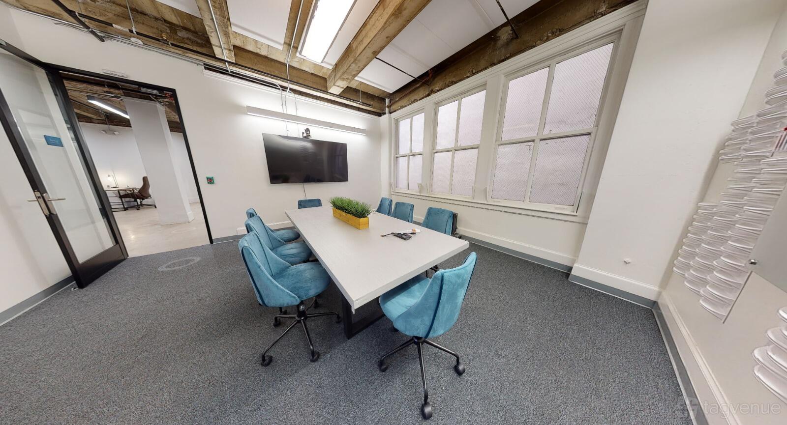 A meeting room with a rectangular table, blue swivel chairs, large windows, and a wall-mounted screen at Dynamico Space.