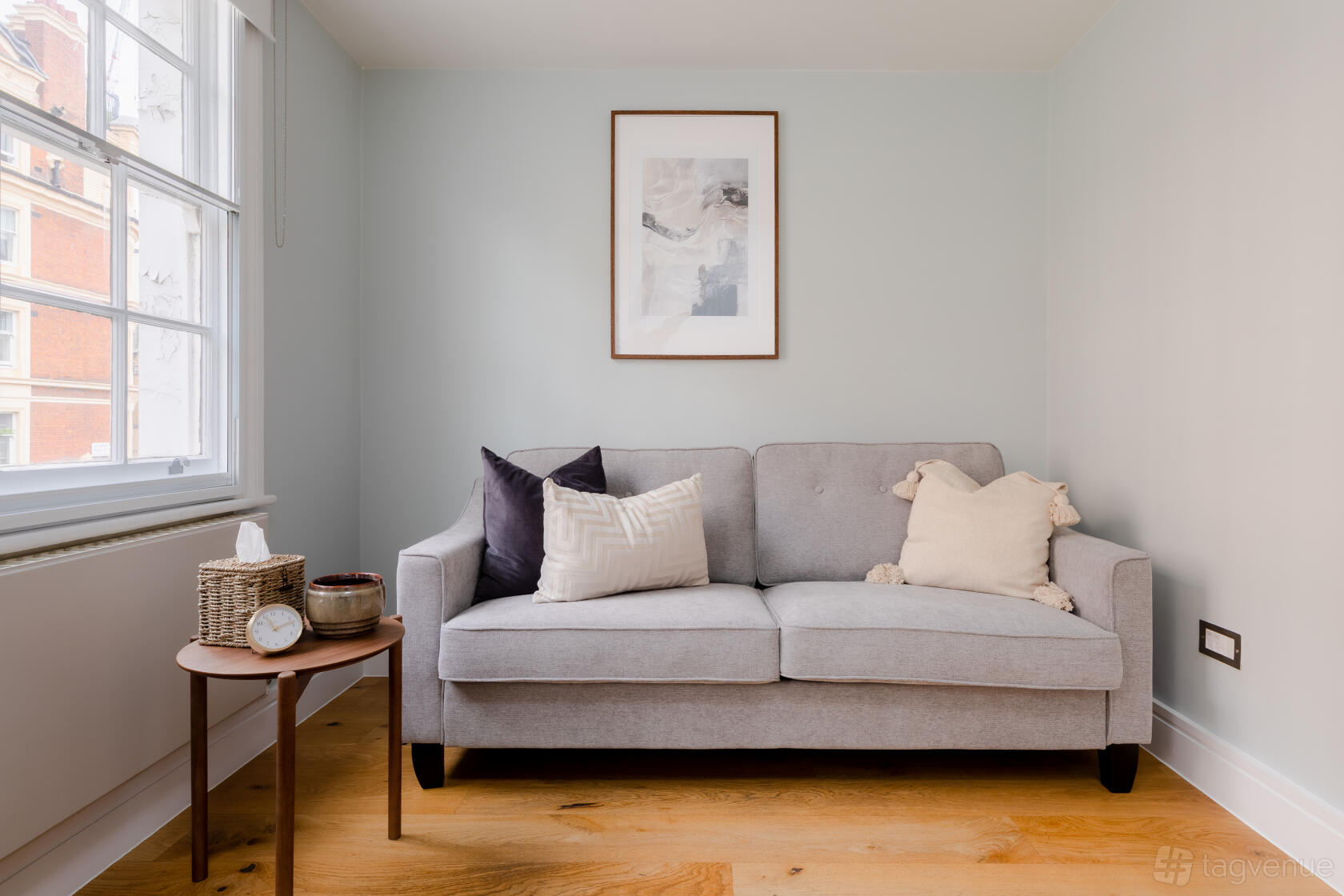 A meeting room with a grey sofa, light wood flooring, a side table, and framed wall art at CuraRooms Marylebone.
