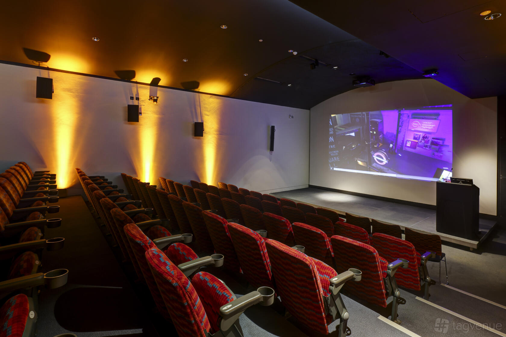 An auditorium with tiered red seats, wall-mounted speakers, and a large projection screen at London Transport Museum.