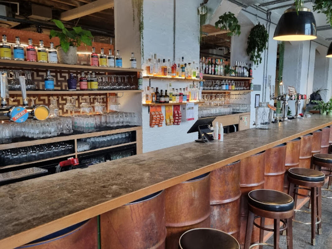A bar with a long wooden counter, shelves of liquor bottles, hanging glasses, and industrial lighting at The Printhouse at Jim & Tonic East.