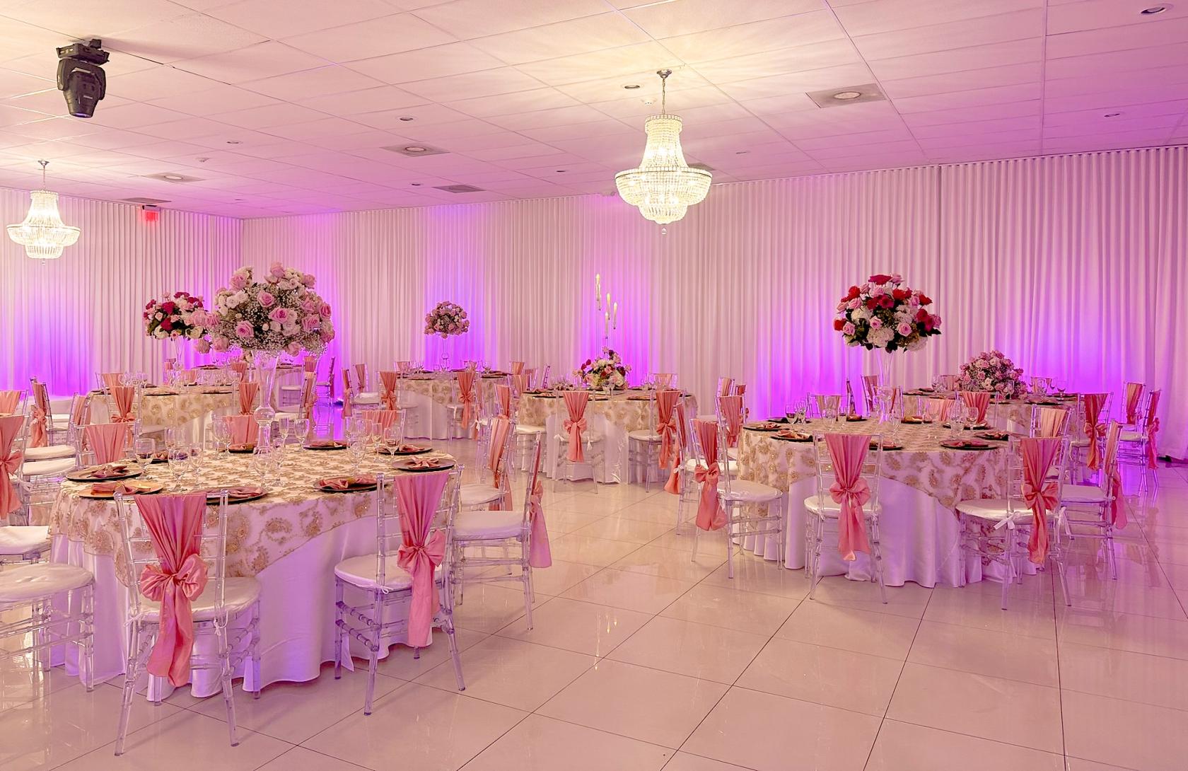 An event space in a hall with chandeliers, round tables dressed in floral linens, pink chair sashes, and uplighting at Forever Reception Hall.