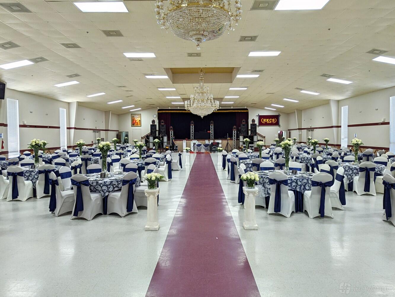 A banquet hall with chandeliers, round tables draped in white linens and navy sashes, at HKCS Community Center.