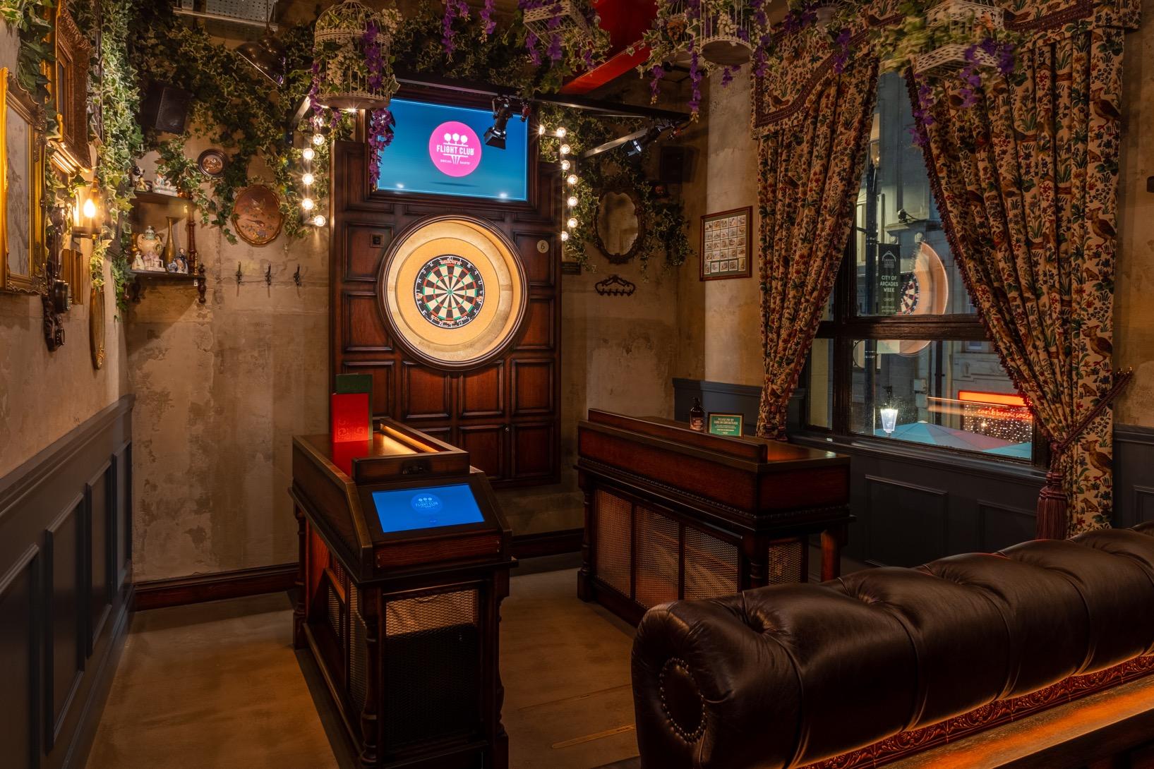 An event space with a dartboard lane, leather seating, floral curtains, and greenery at Flight Club Cardiff.