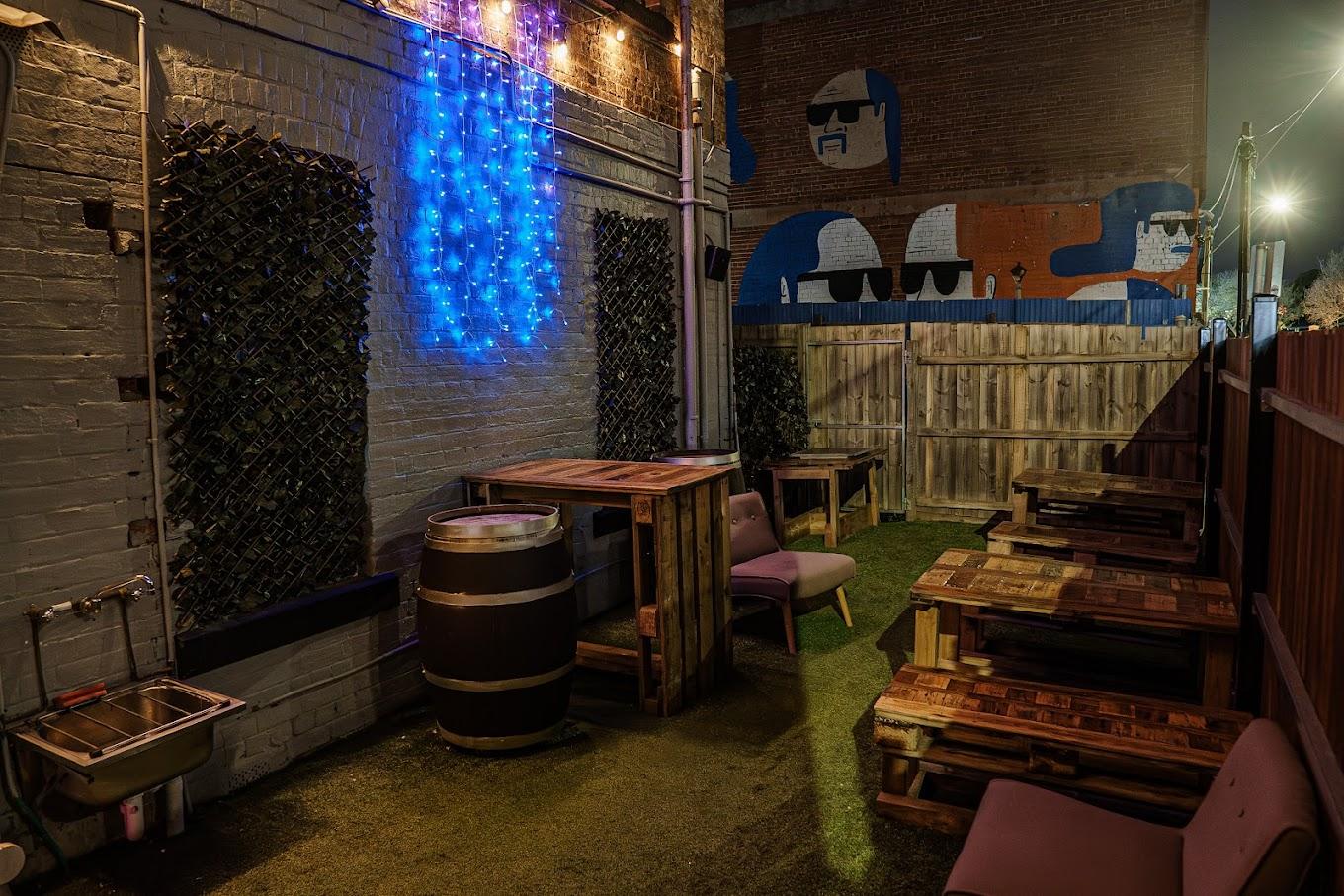 An outdoor bar area with pallet tables, barrel seating, artificial grass, and blue string lights at XO Bar.