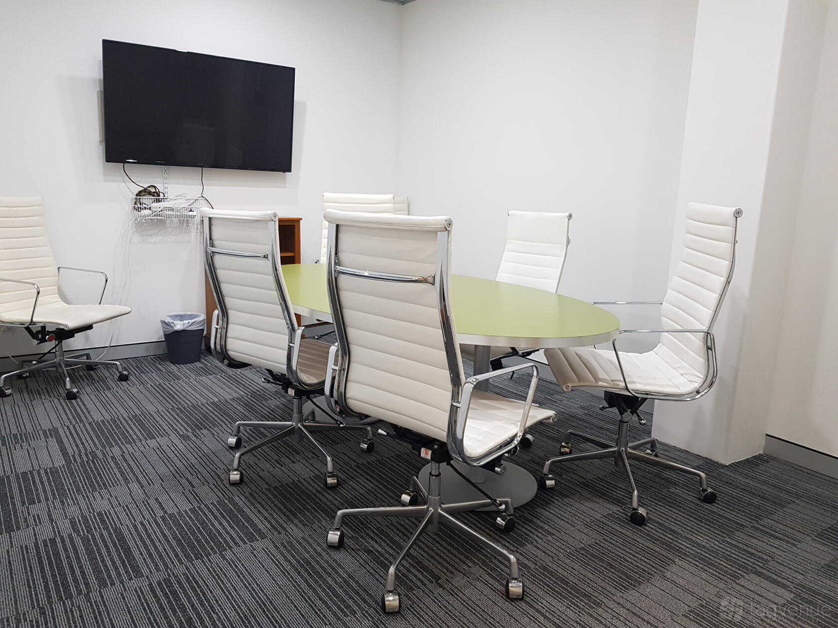 A meeting room with a glass-topped oval table, white leather chairs, and wall-mounted TV at A23.