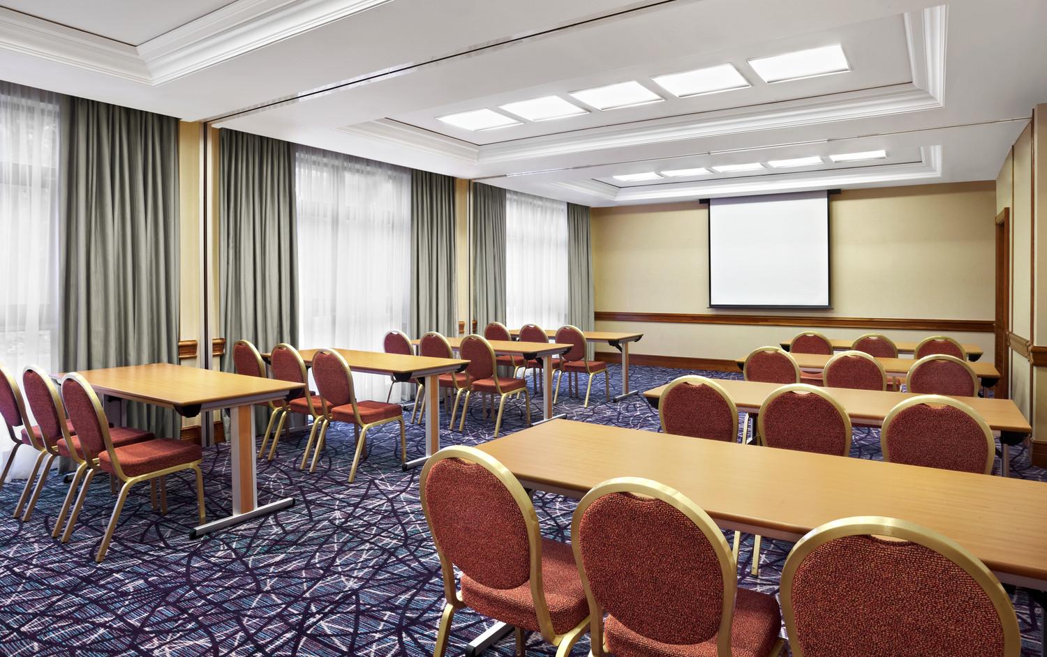 A meeting room with rows of red cushioned chairs, rectangular tables, large windows, and a projector screen at Sheraton Skyline Hotel London Heathrow.