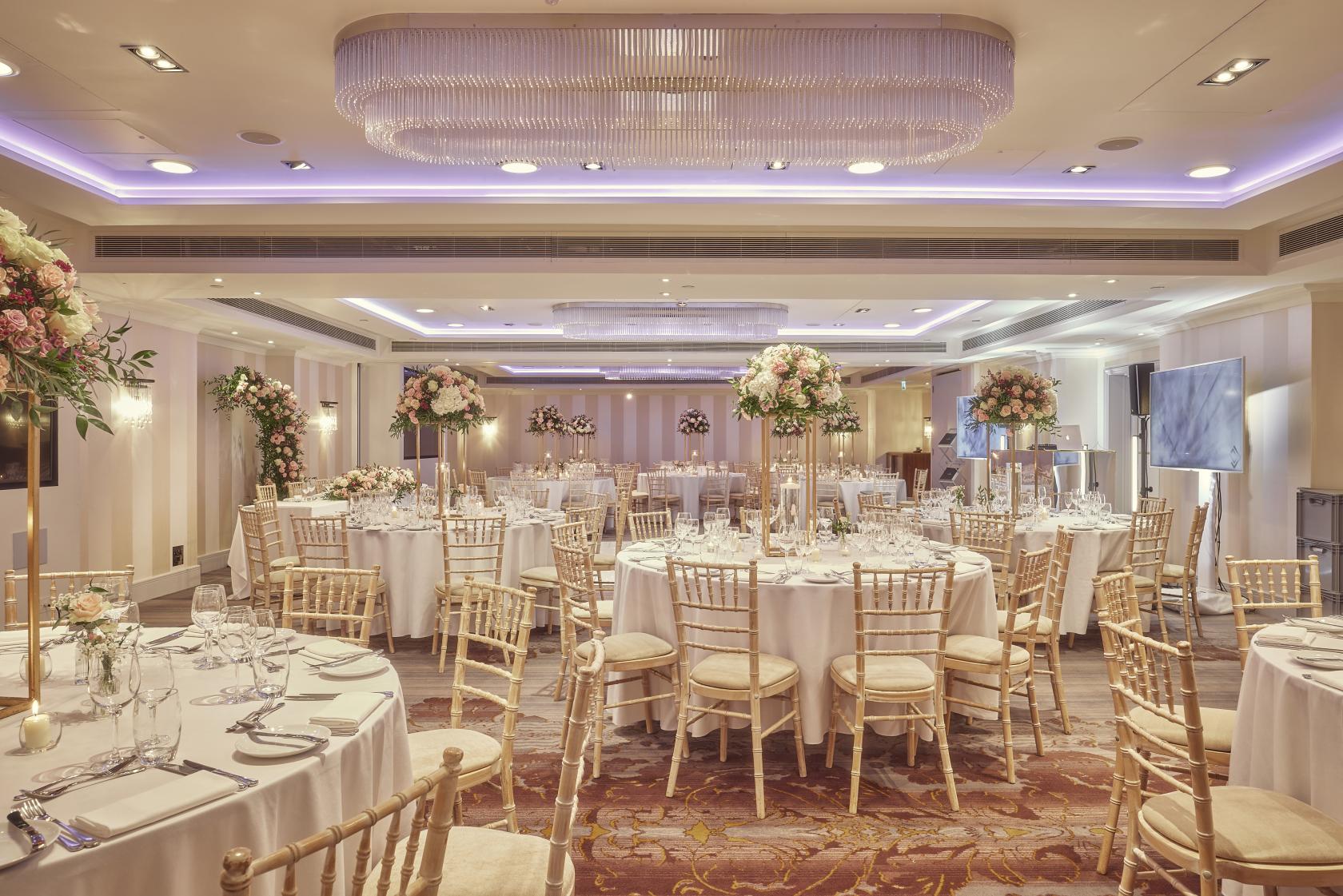A hotel ballroom with round tables, white linens, gold chairs, and tall floral centerpieces at Meetings & Events Venues.