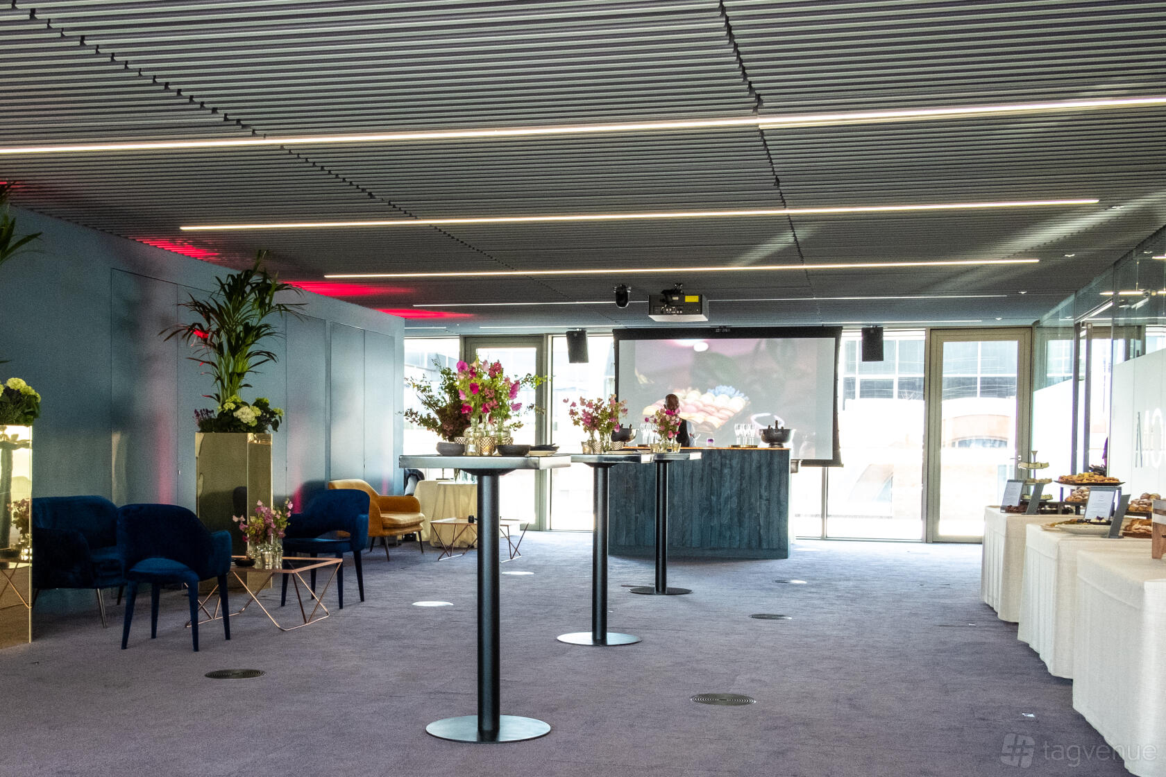 A function room with floor-to-ceiling windows, lounge seating, and high tables with floral centerpieces at Events at No6.