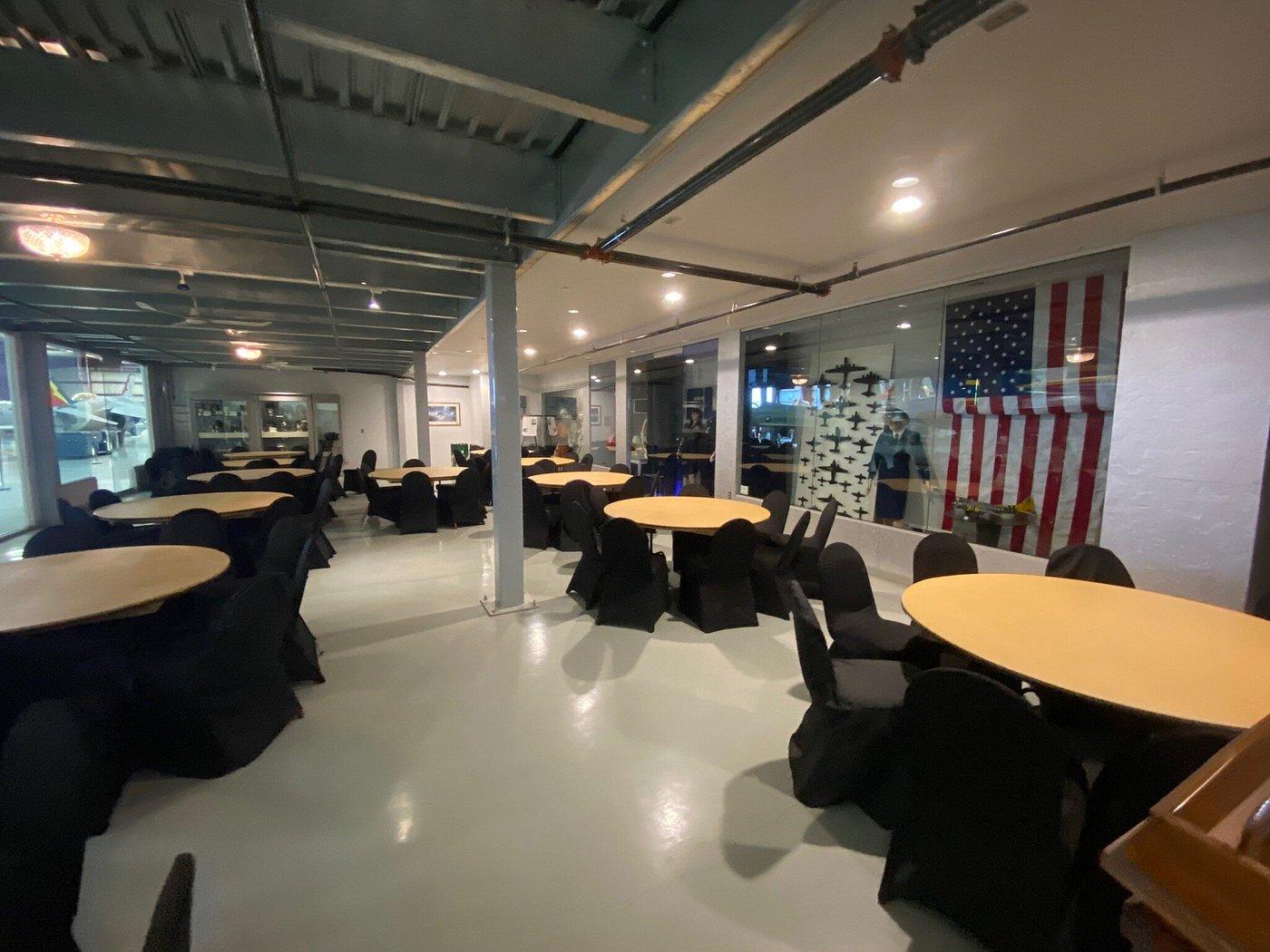 An event space in a museum with round tables, black chairs, and aviation displays beside an American flag at Yanks Air Museum.