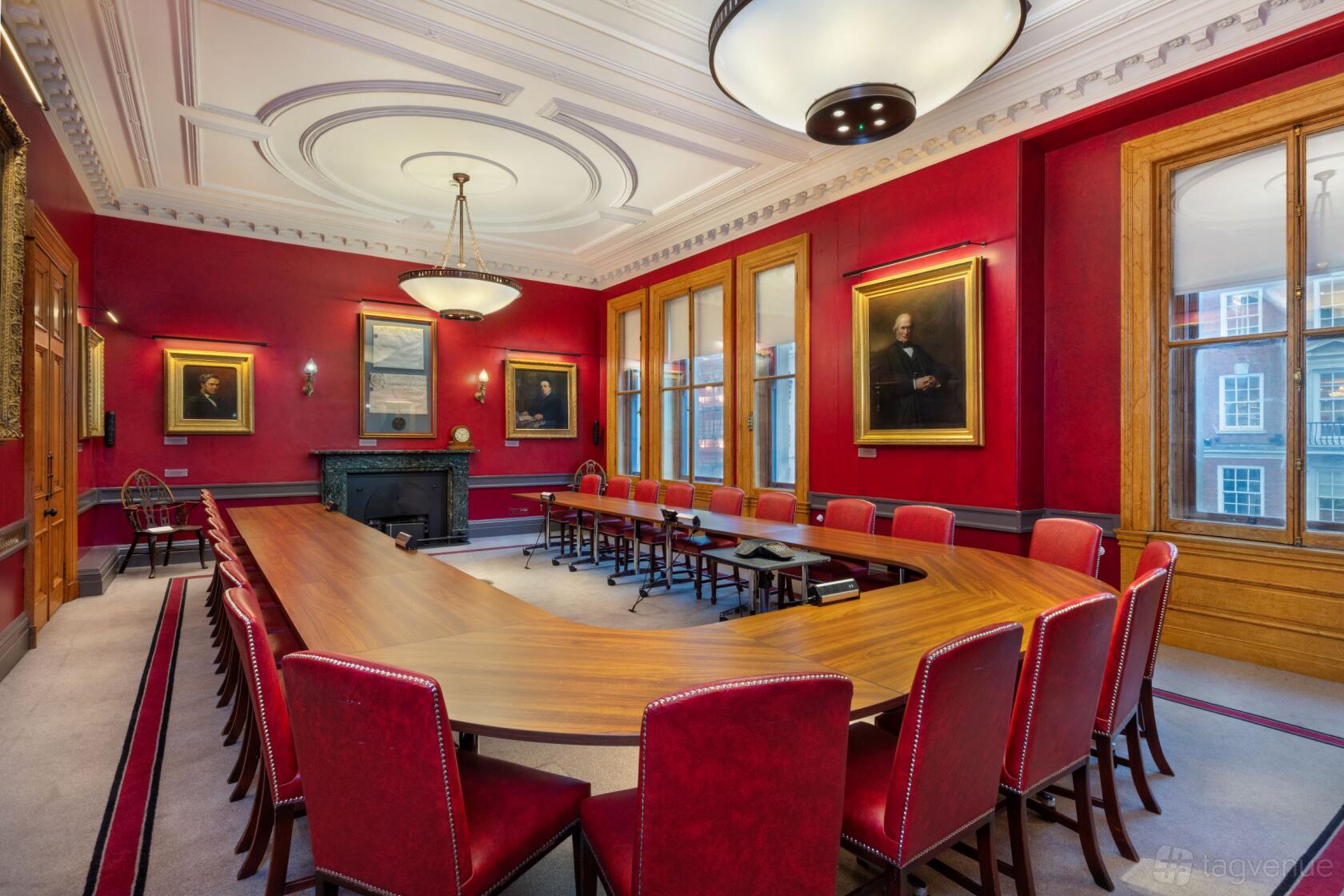 A formal meeting room with red walls, portraits, a U-shaped wooden table, and large windows at The Geological Society.