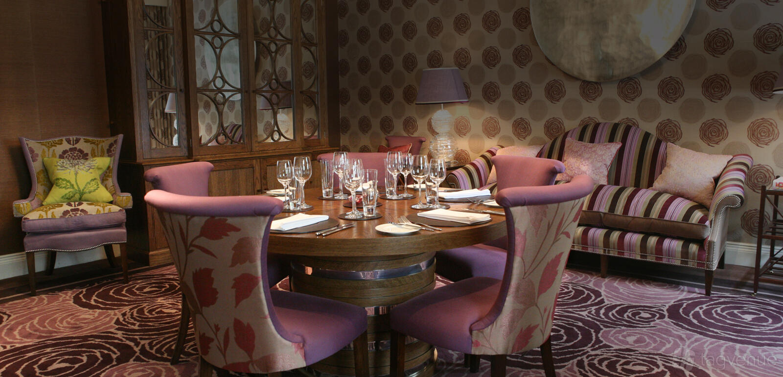 A hotel lounge with patterned lilac chairs, a round wooden table set for dining, and floral wallpaper at The Roseate Reading.