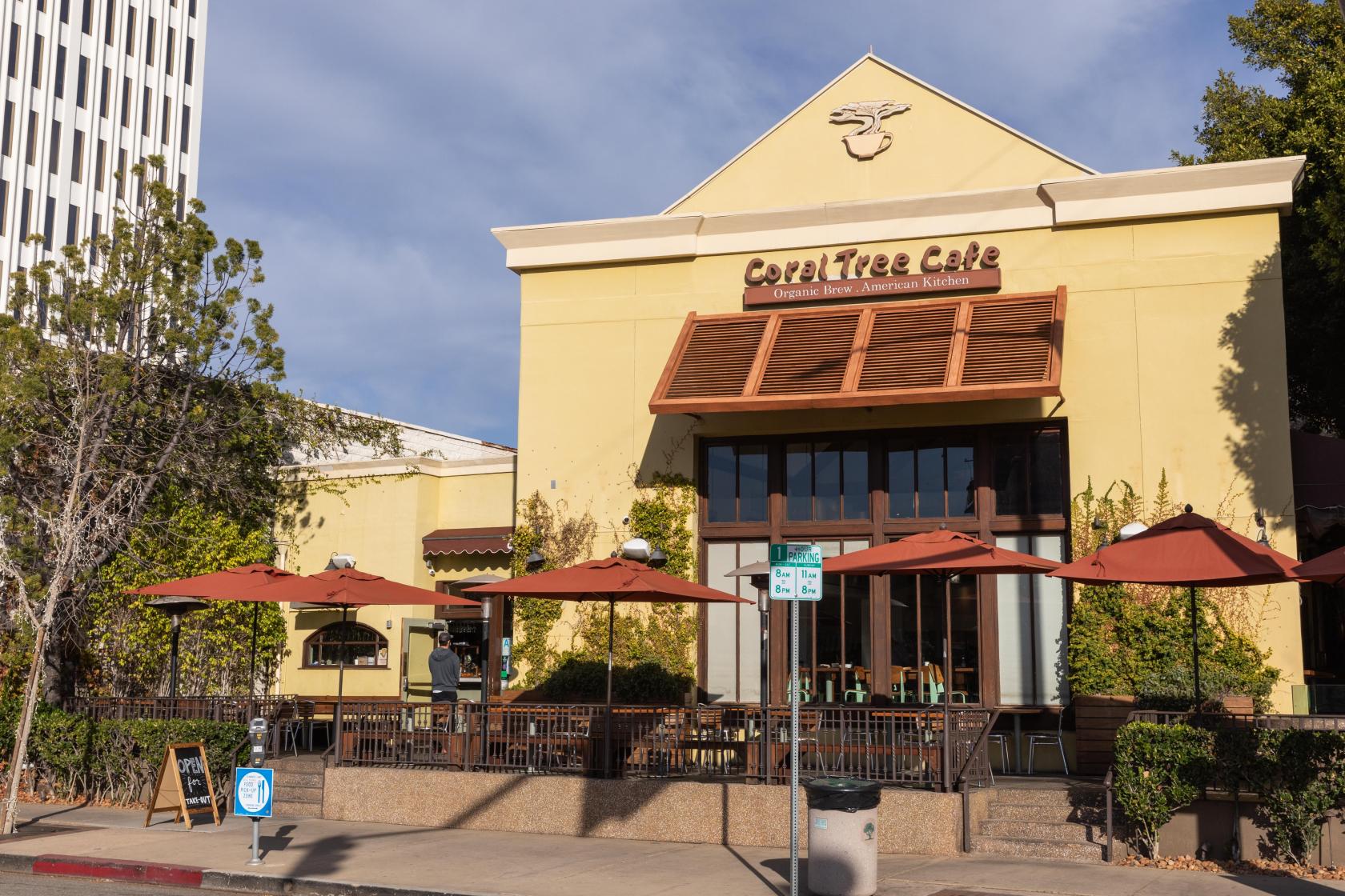 An outdoor restaurant patio with red umbrellas, vine-covered railings, and sidewalk seating at Coral Tree Cafe - Brentwood.