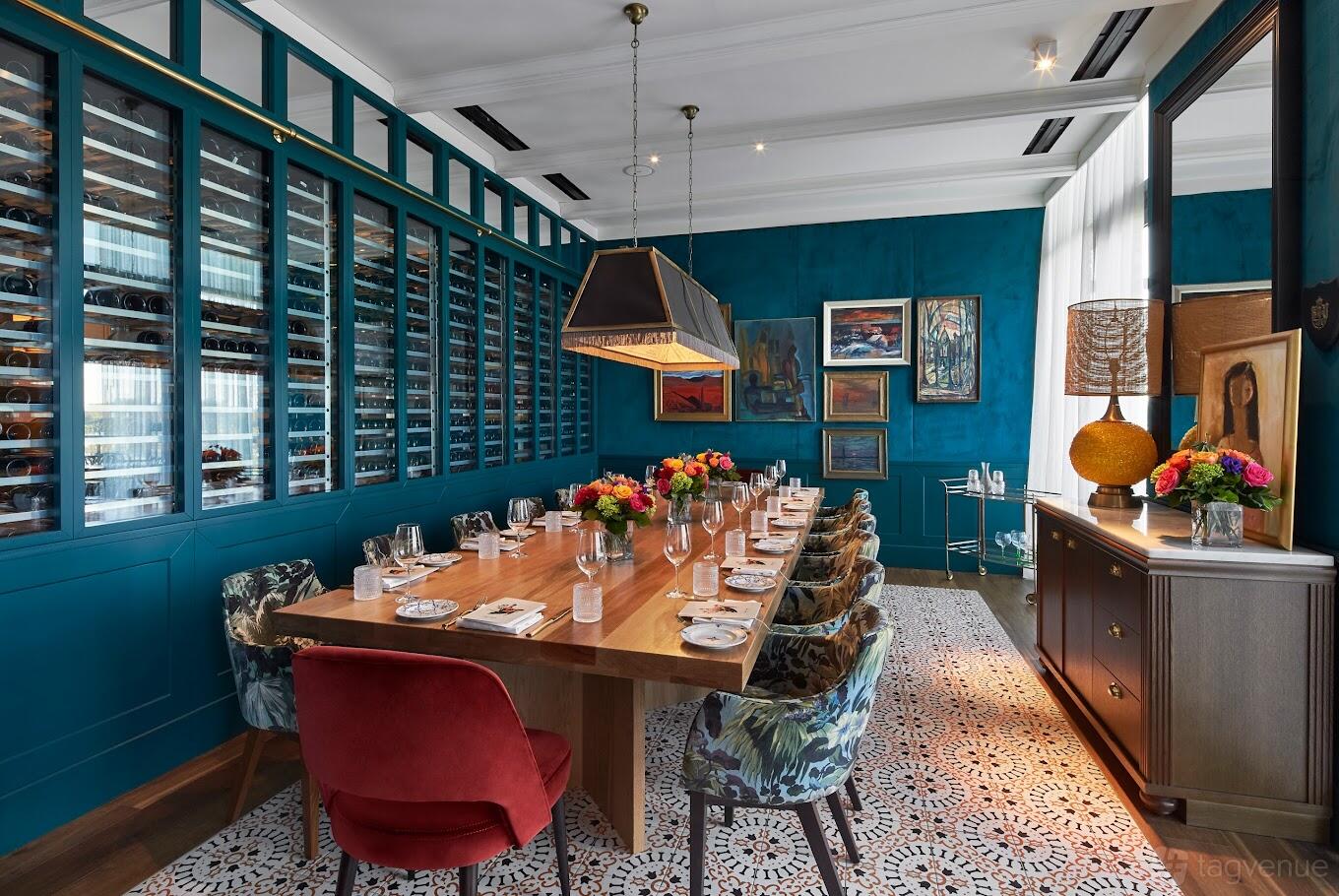 A dining room with a long wooden table, patterned tile floor, colorful chairs, and a glass wine wall at Del Mar.