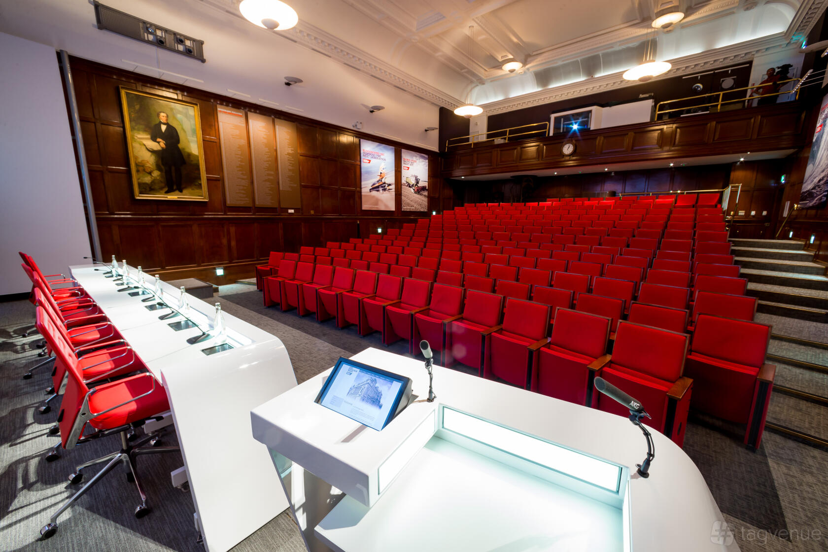 An auditorium with red tiered seating, wood-paneled walls, and a panelist table with microphones at One Birdcage Walk.