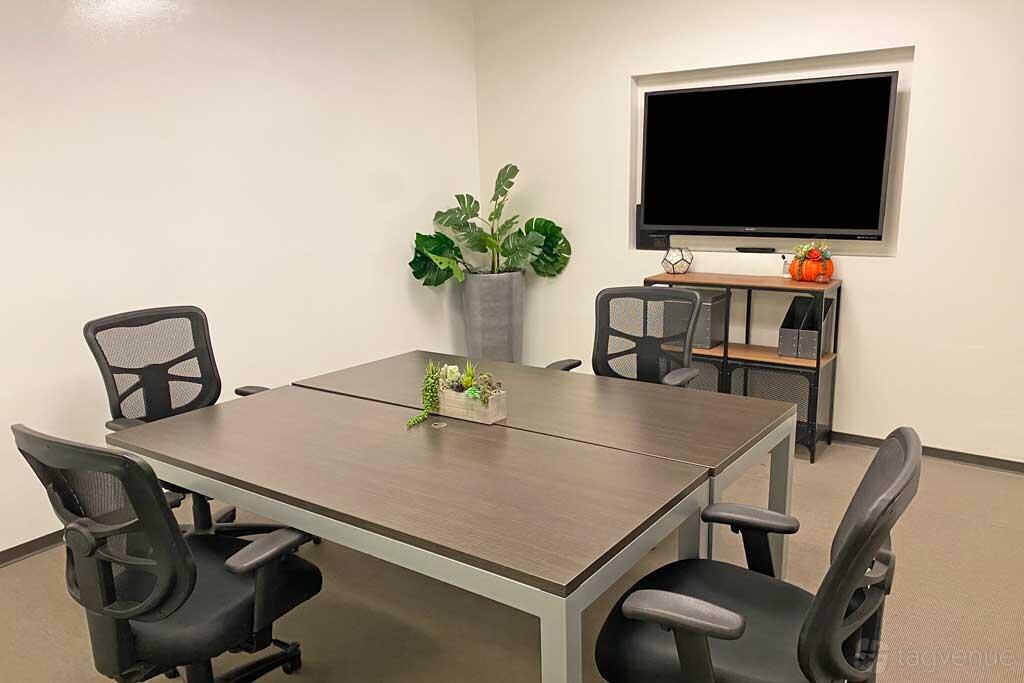 A coworking meeting room with a square table, mesh office chairs, a wall-mounted screen, and potted plants at Premier Workspaces - 4590 MacArthur.
