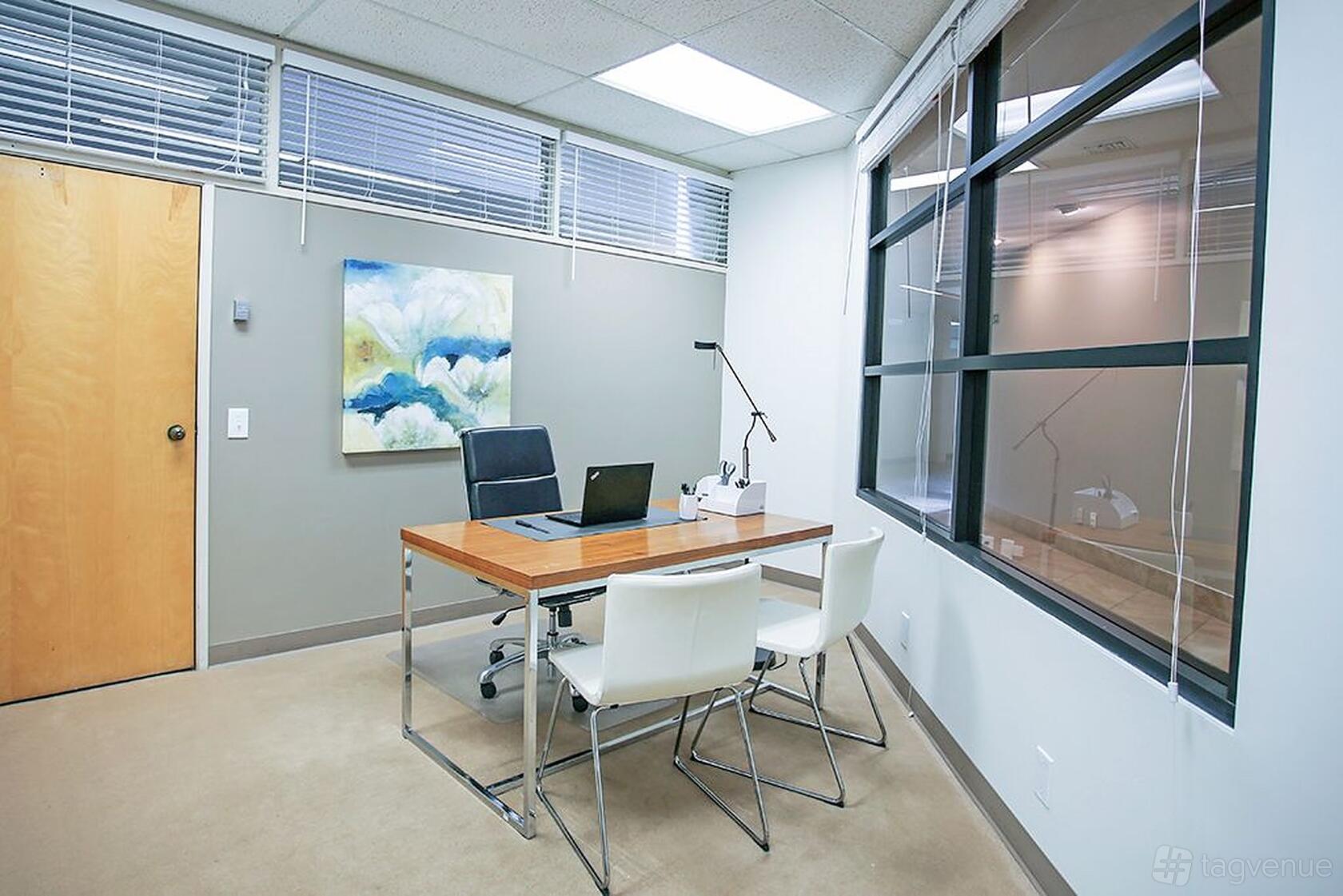 A coworking space with a wooden desk, two white chairs, and large interior windows at Premier Workspaces - Airport Executive Suites.
