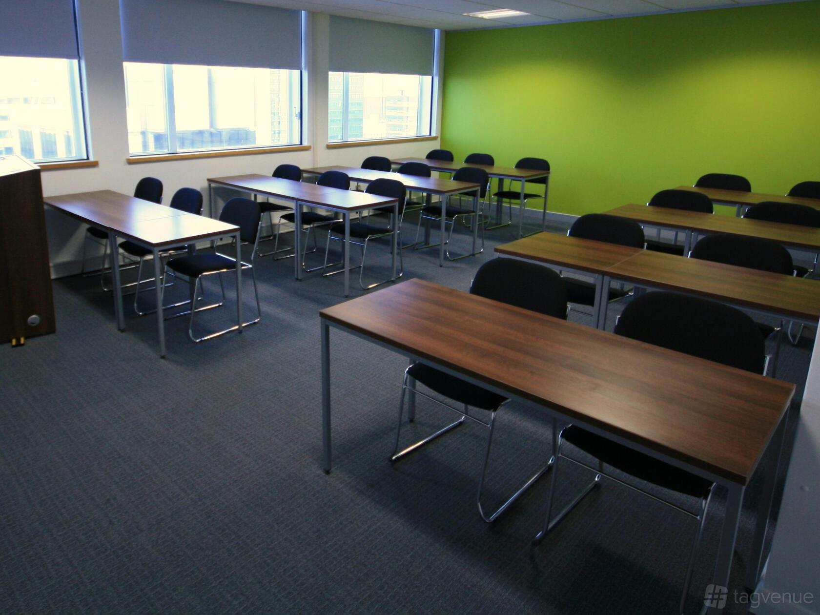 A meeting centre room with rows of wooden tables, black chairs, large windows, and a green accent wall at First Intuition Manchester.