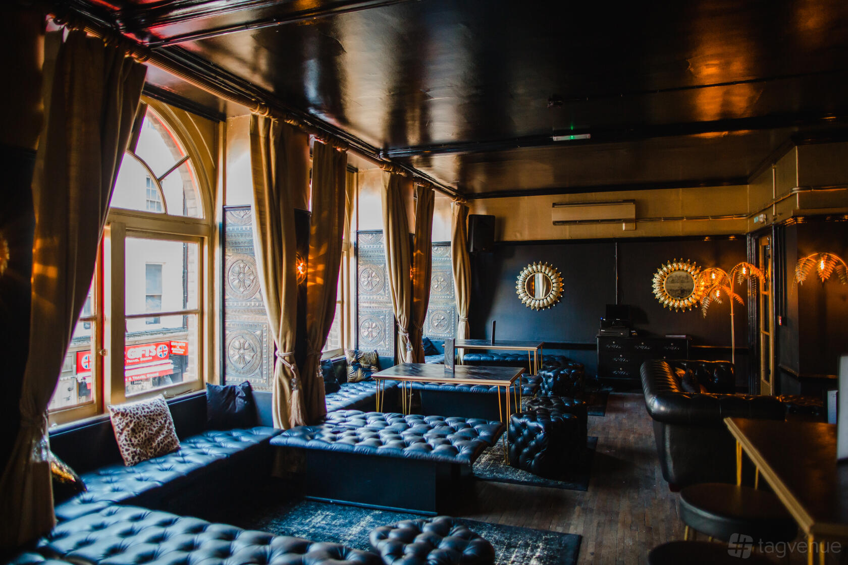 A cocktail bar with tufted black leather seating, large arched windows, and gold-framed mirrors at Covent Garden Social Club.