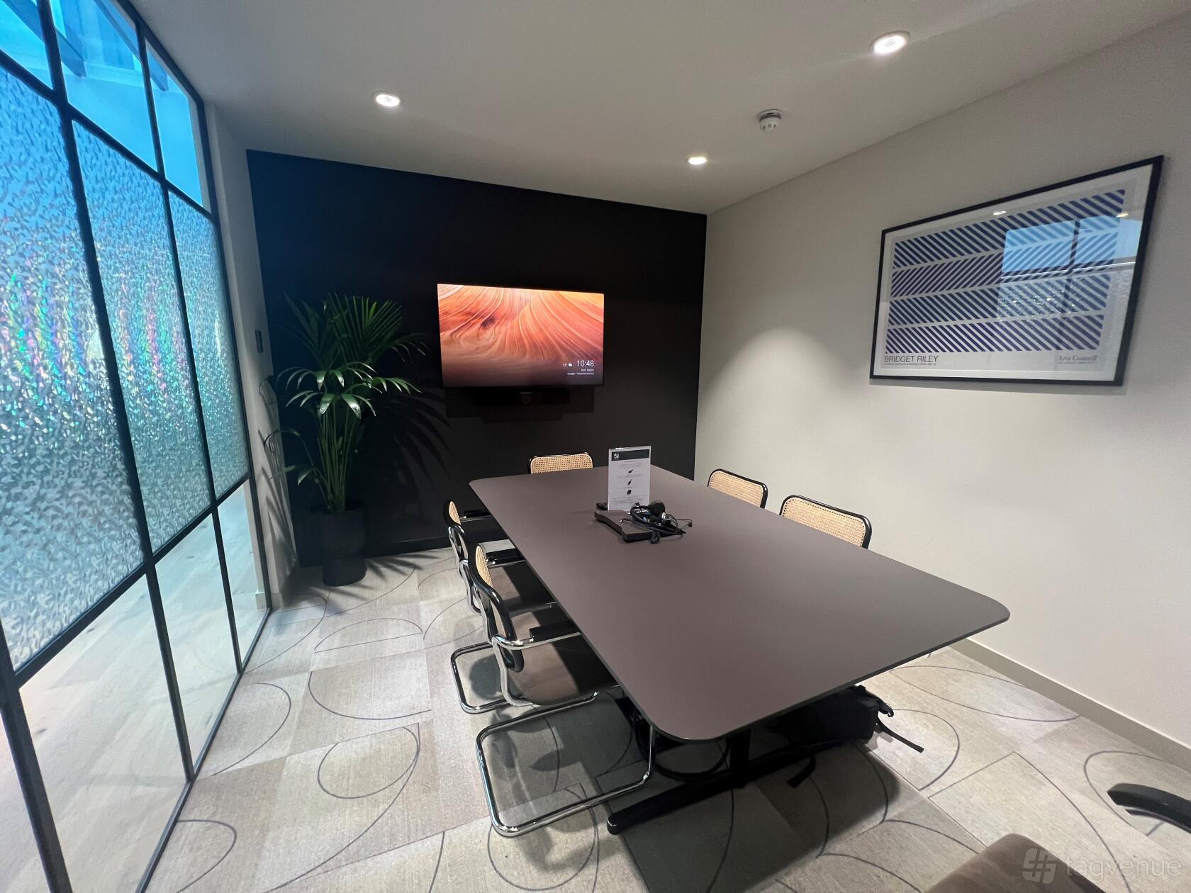A meeting room with a large black conference table, six chairs, wall-mounted screen, and glass partition at Phoenix House.