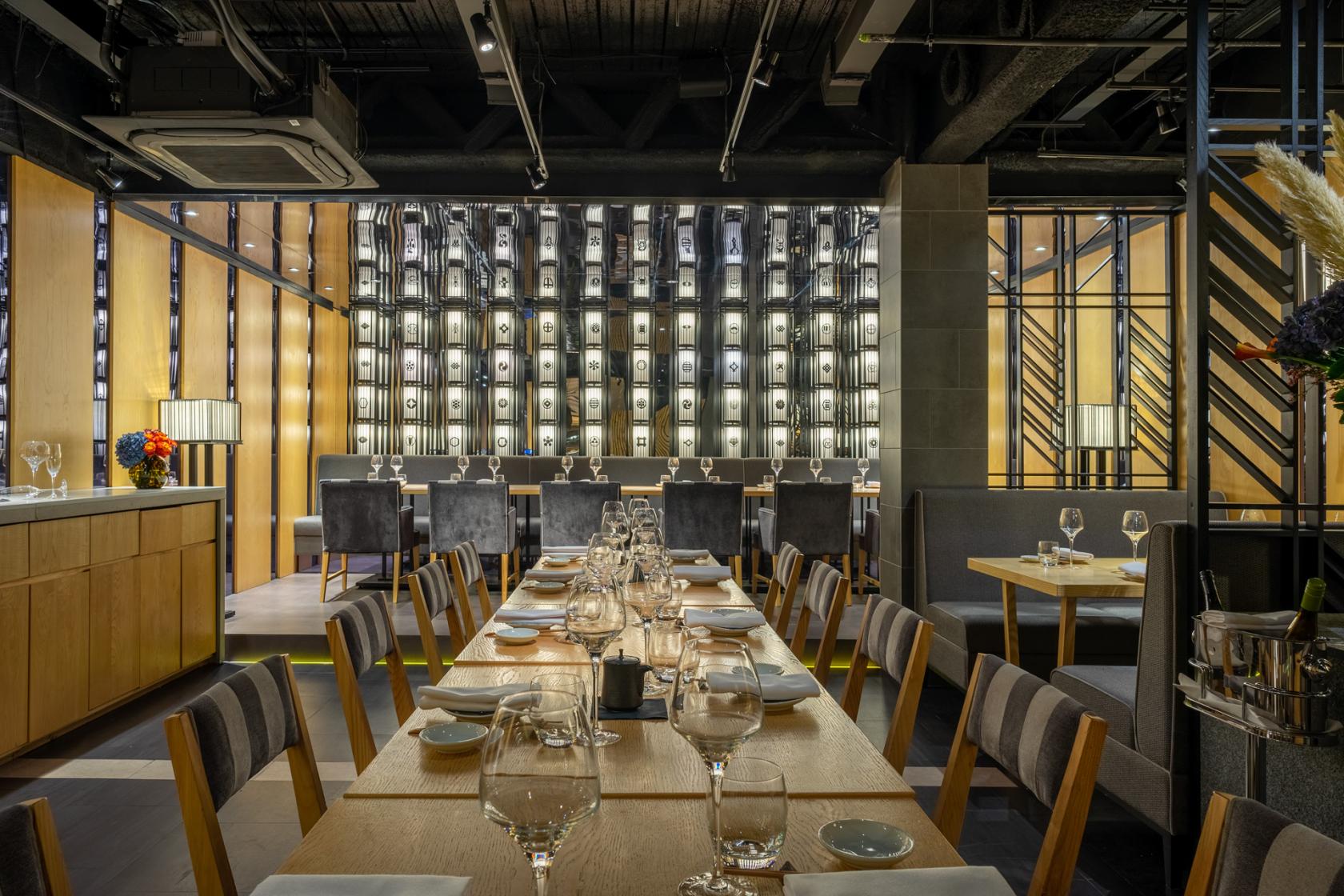 A restaurant dining area with a long wooden table, cushioned chairs, and a backlit wall of decorative bottles at Ginza St. James's.