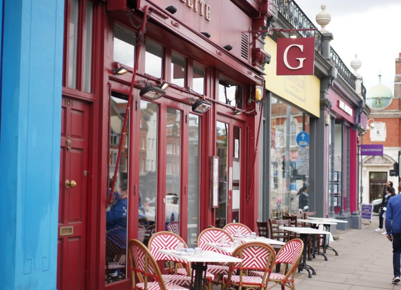 A restaurant with red-framed windows and outdoor seating with white and red chairs at Gazette Putney.
