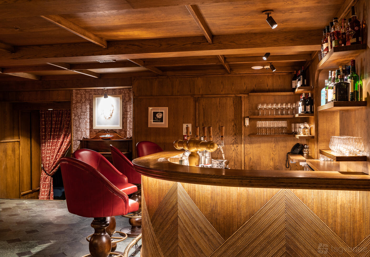 A pub bar with a wooden counter, brass beer taps, red upholstered stools, and wood-paneled walls at The Coach Makers Arms Pub Marylebone.