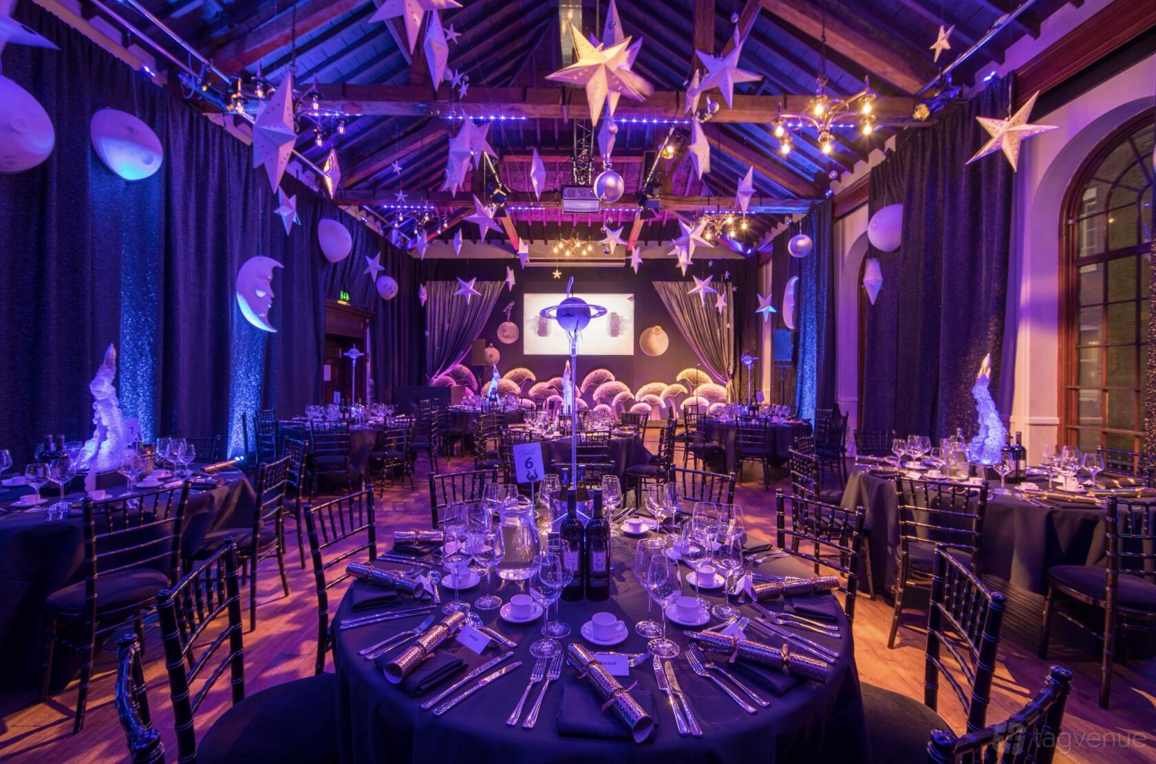 An event space with high timber ceilings, purple mood lighting, and round tables set for dinner at The Brewery.