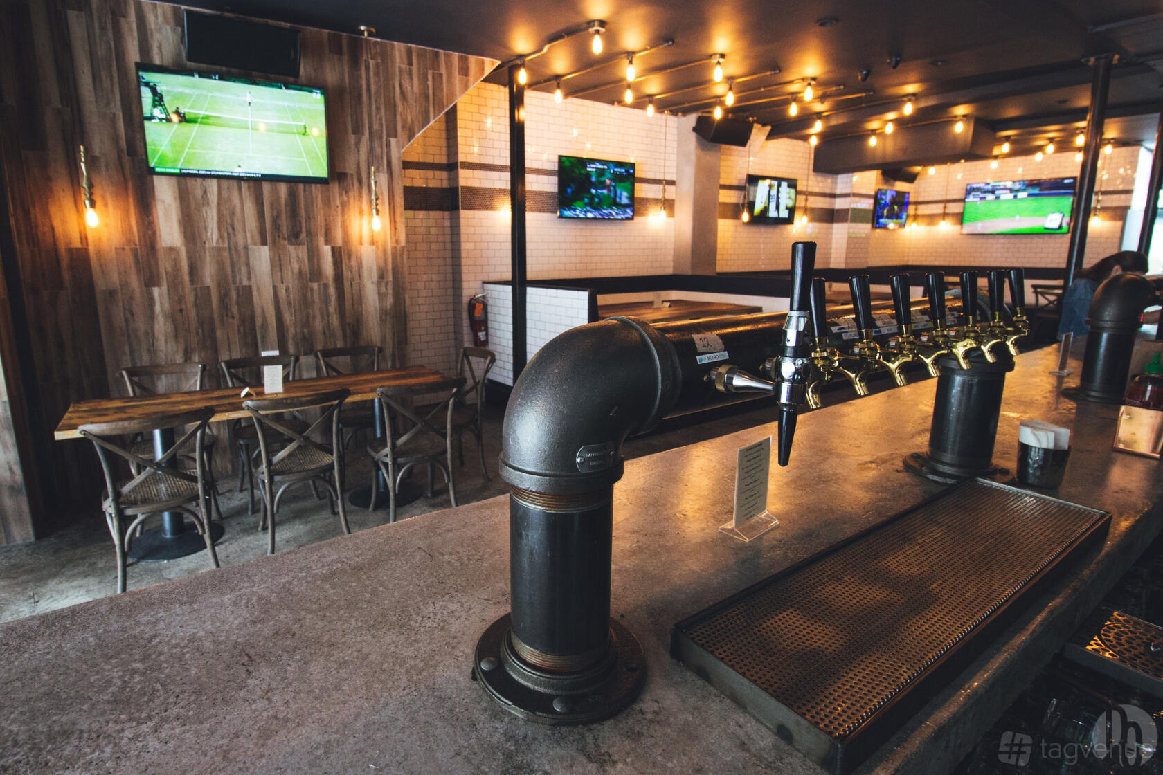An event space in a bar with a long tap system, wood furniture, wall-mounted TVs, and exposed bulbs at Emblem Sports Bar.