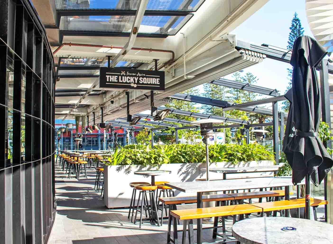 An outdoor bar area with long wooden tables, bench seating, and large planters beneath a retractable roof at The Lucky Squire.