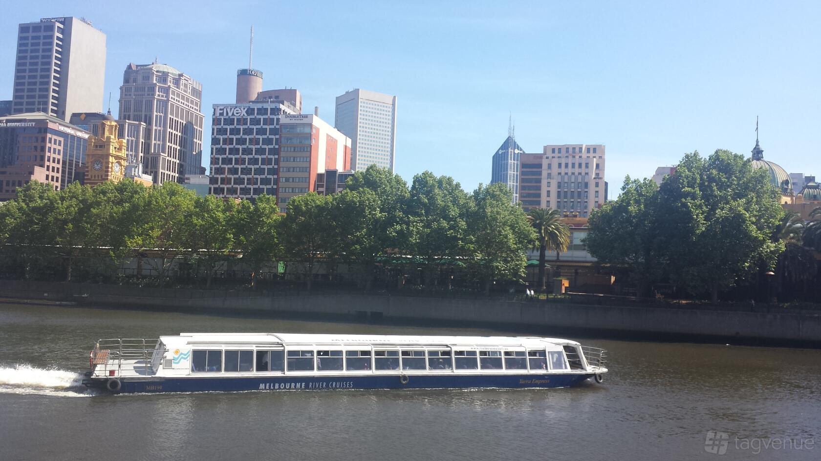 A river boat with glass windows and covered seating cruising past city buildings on the Yarra Empress.