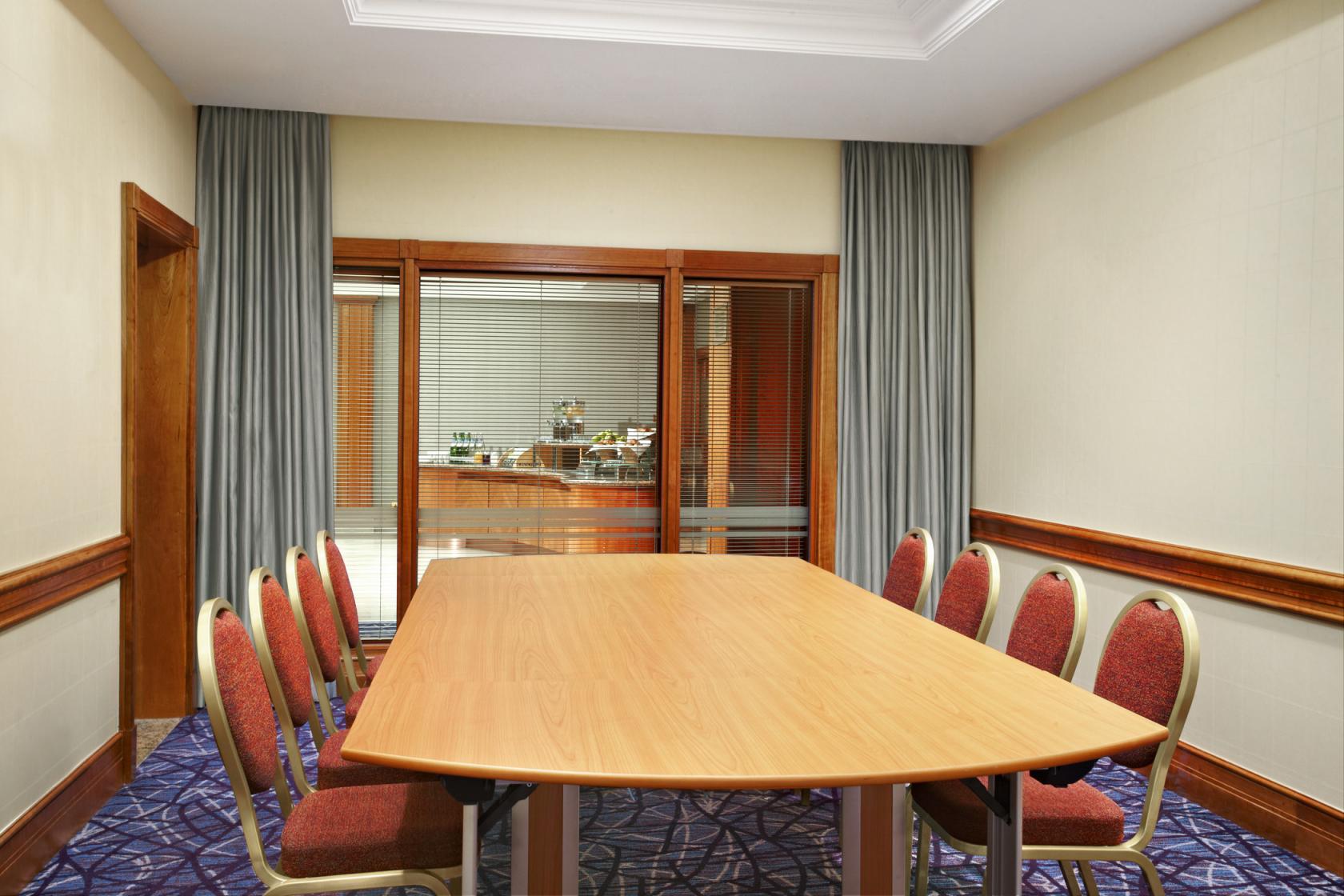 A meeting room with a long wooden table, red upholstered chairs, and floor-to-ceiling curtains at Sheraton Skyline Hotel London Heathrow.