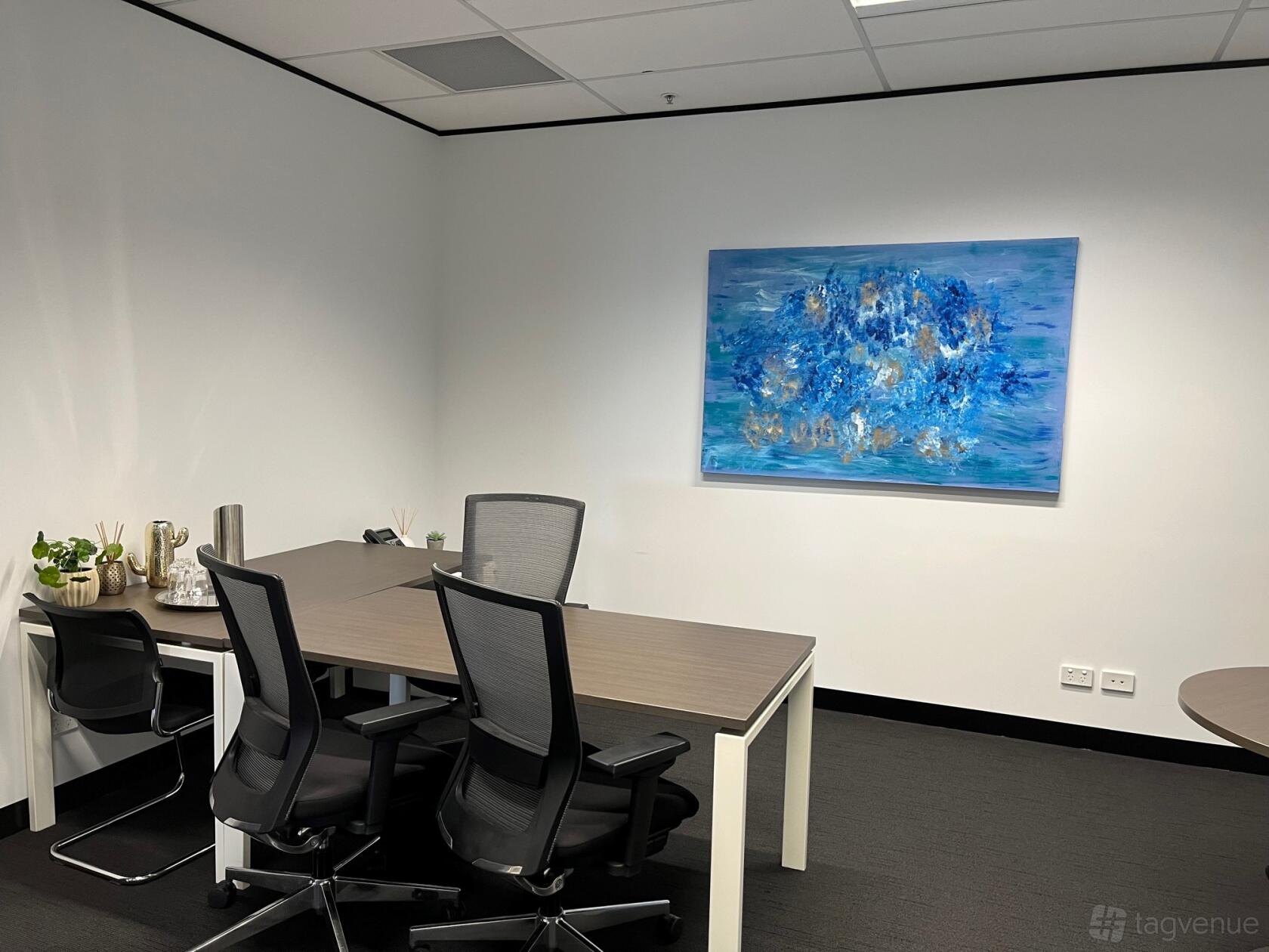 A meeting room with two desks, black ergonomic chairs, and a blue abstract painting at The Work Studio.